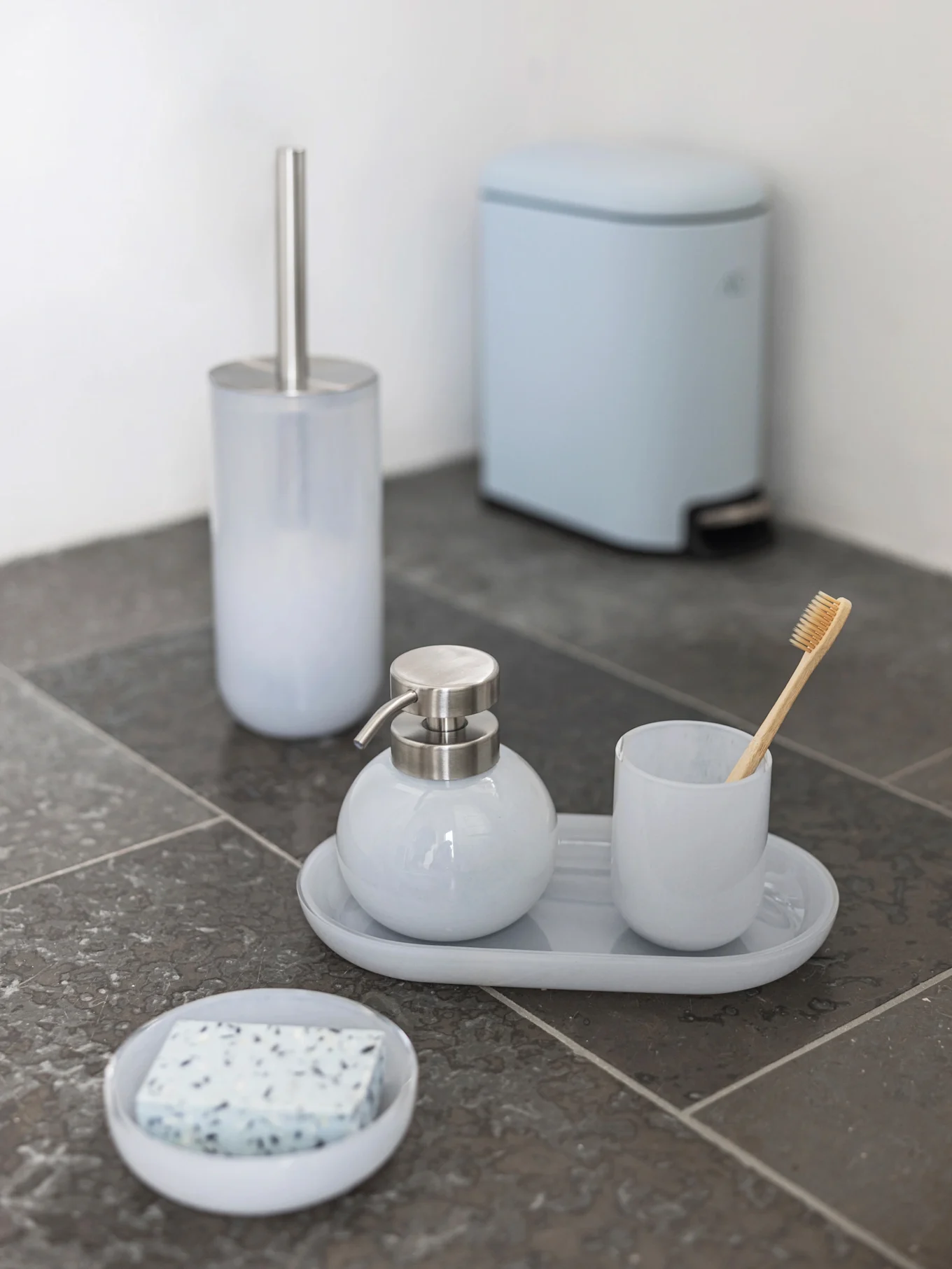 Set bagno azzurro: scopino, dispenser sapone, bicchiere con spazzolino di bambù, portasapone e pattumiera a pedale su piastrelle scure.