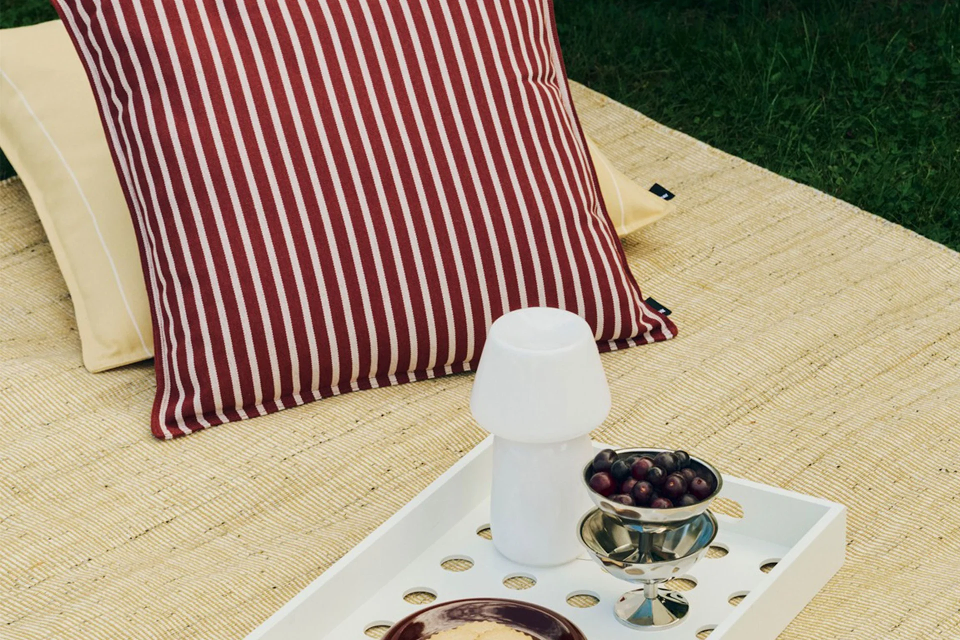 Allestimento da picnic all'aperto con cuscino a righe rosse e bianche, cuscino giallo, vassoio bianco con lampada a fungo e una ciotola di bacche.