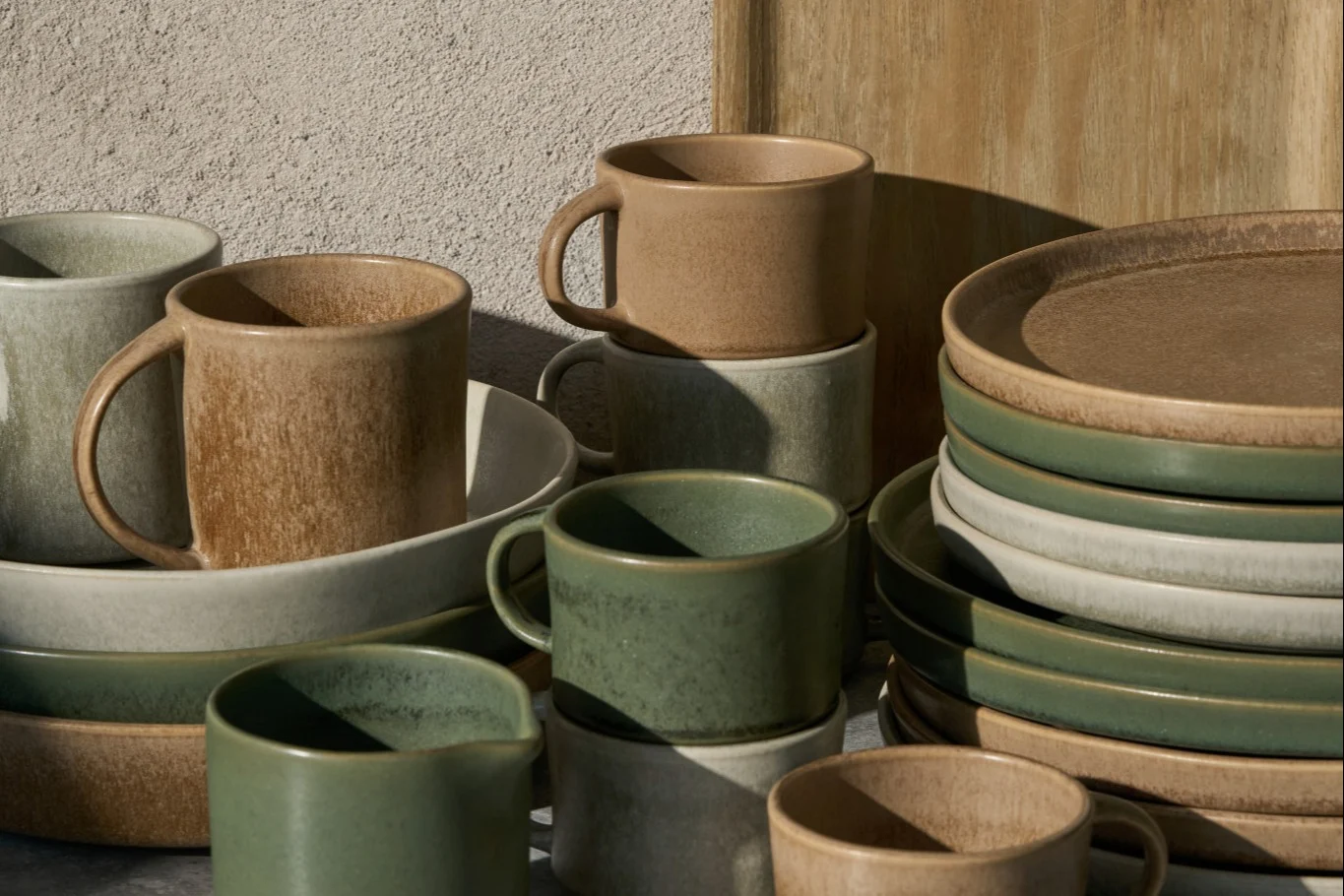 Tazze, ciotole e piatti in ceramica impilati in beige, grigio chiaro e verde salvia, con finitura rustica.