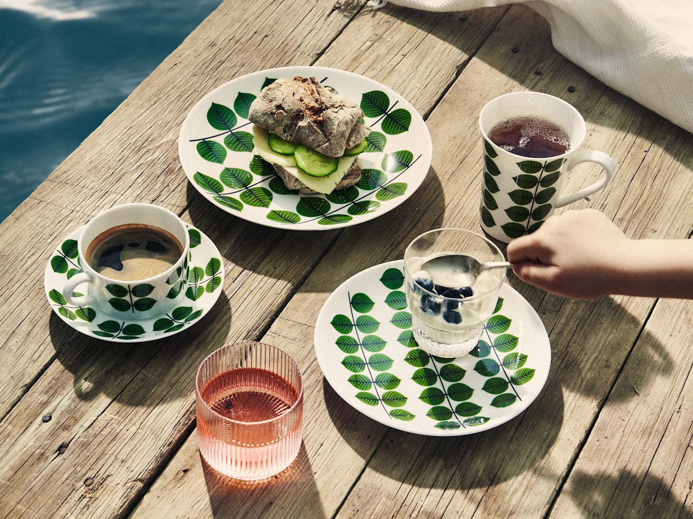 Colazione su molo di legno: piatti a foglie, sandwich, caffè, tè, yogurt con mirtilli e bevanda rosa.