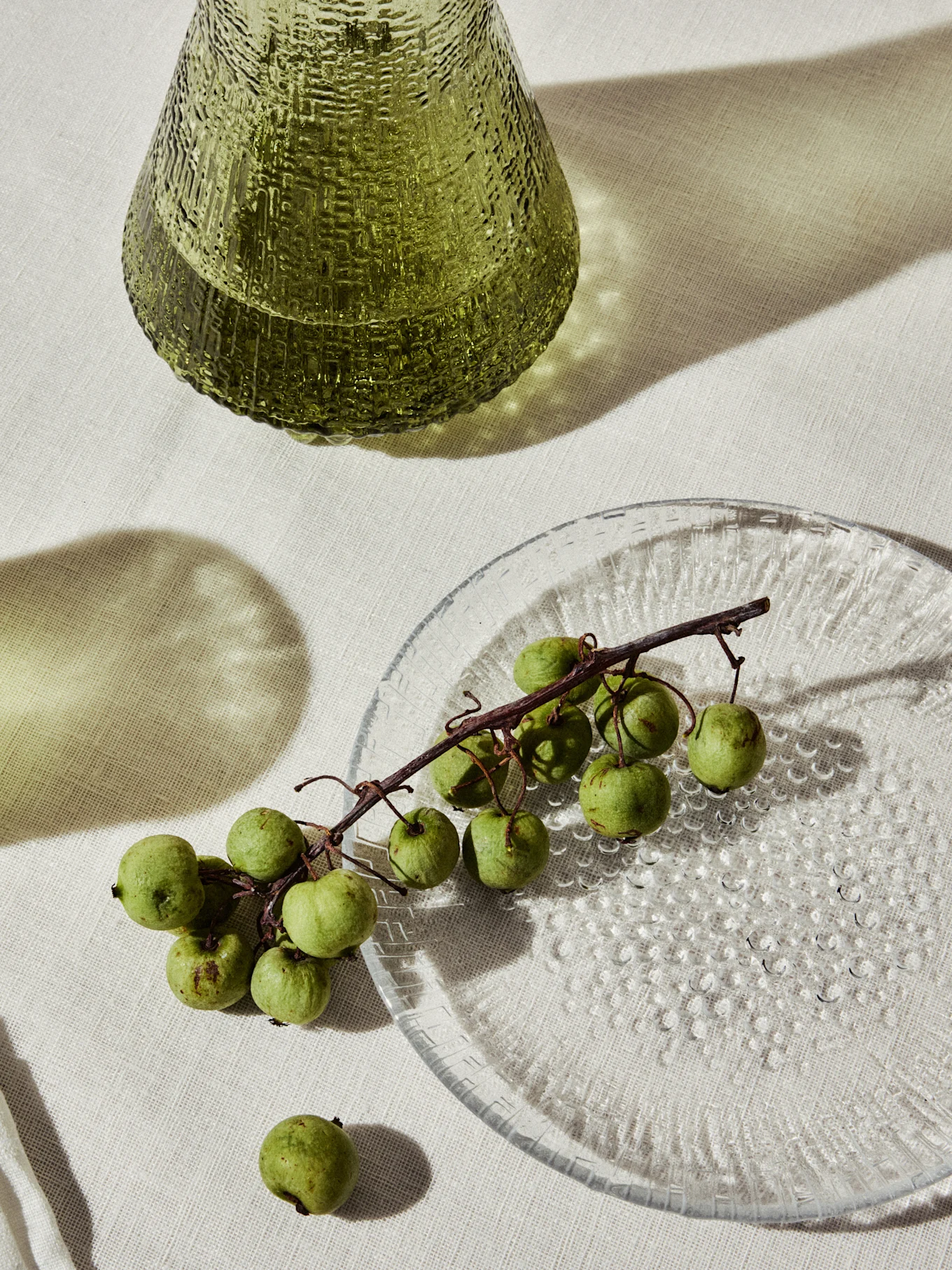 Vaso in vetro verde testurizzato e un piatto trasparente con piccoli frutti verdi su un ramo.