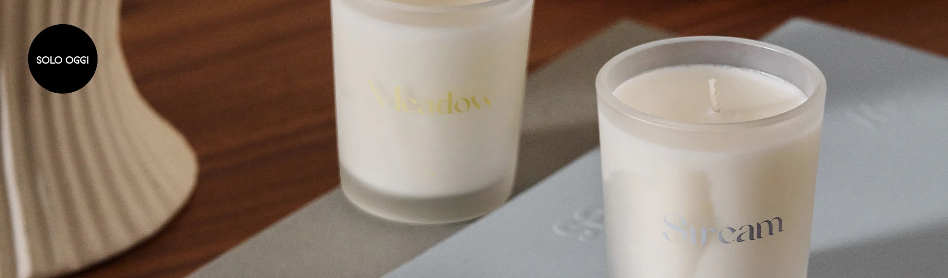 Due candele bianche in barattoli di vetro satinato, una etichettata 'Meadow' e l'altra 'Stream', con logo 'SOLO OGGI'.
