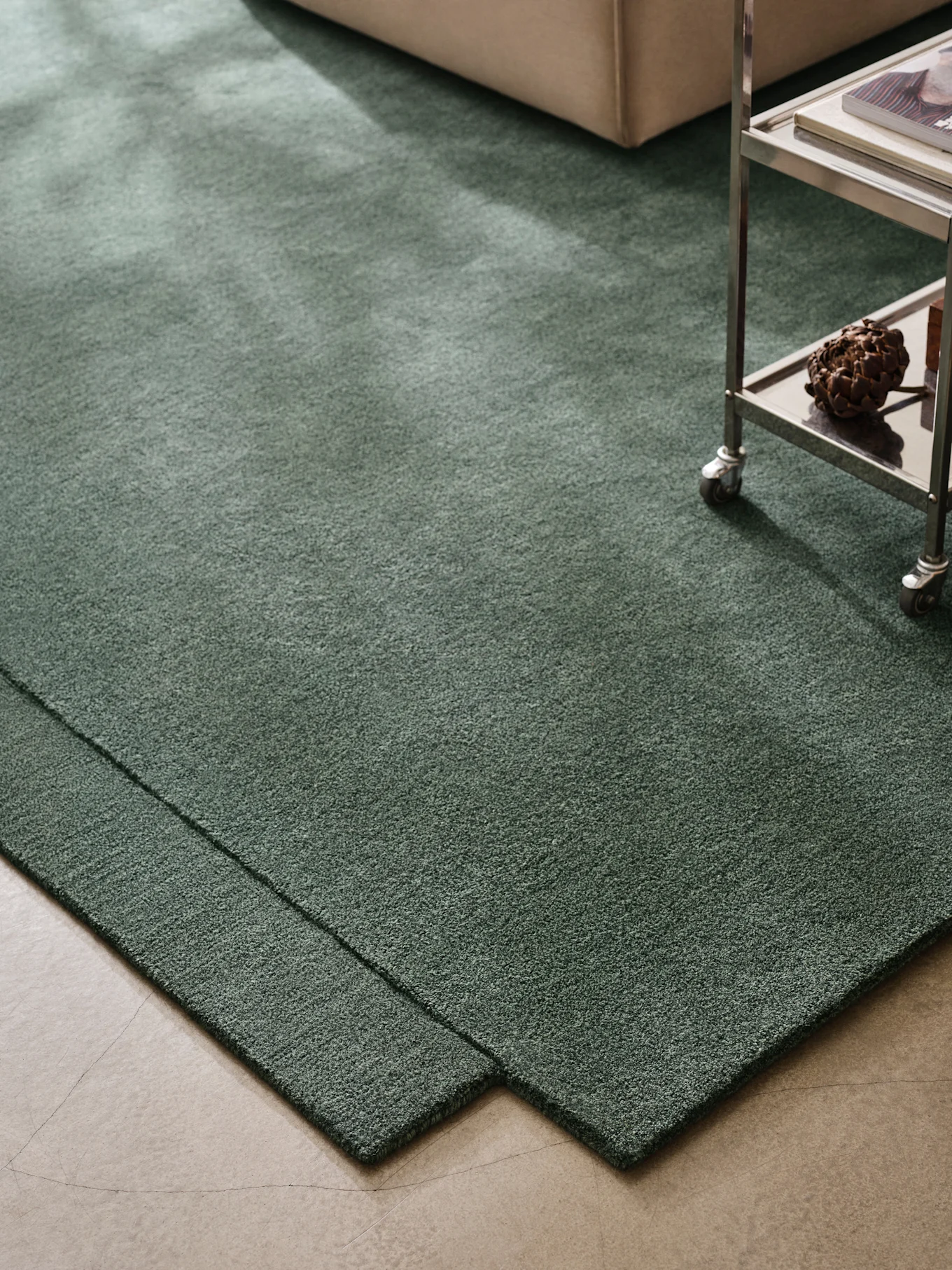 Tappeto shaggy verde scuro su pavimento in cemento, con divano beige e carrello in metallo.