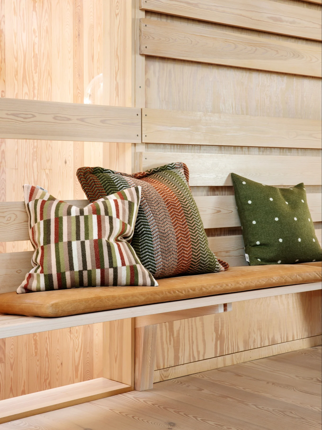 Tre cuscini decorativi su una panca di legno con cuscino in pelle. Uno a righe, uno a spina di pesce e uno verde a pois.