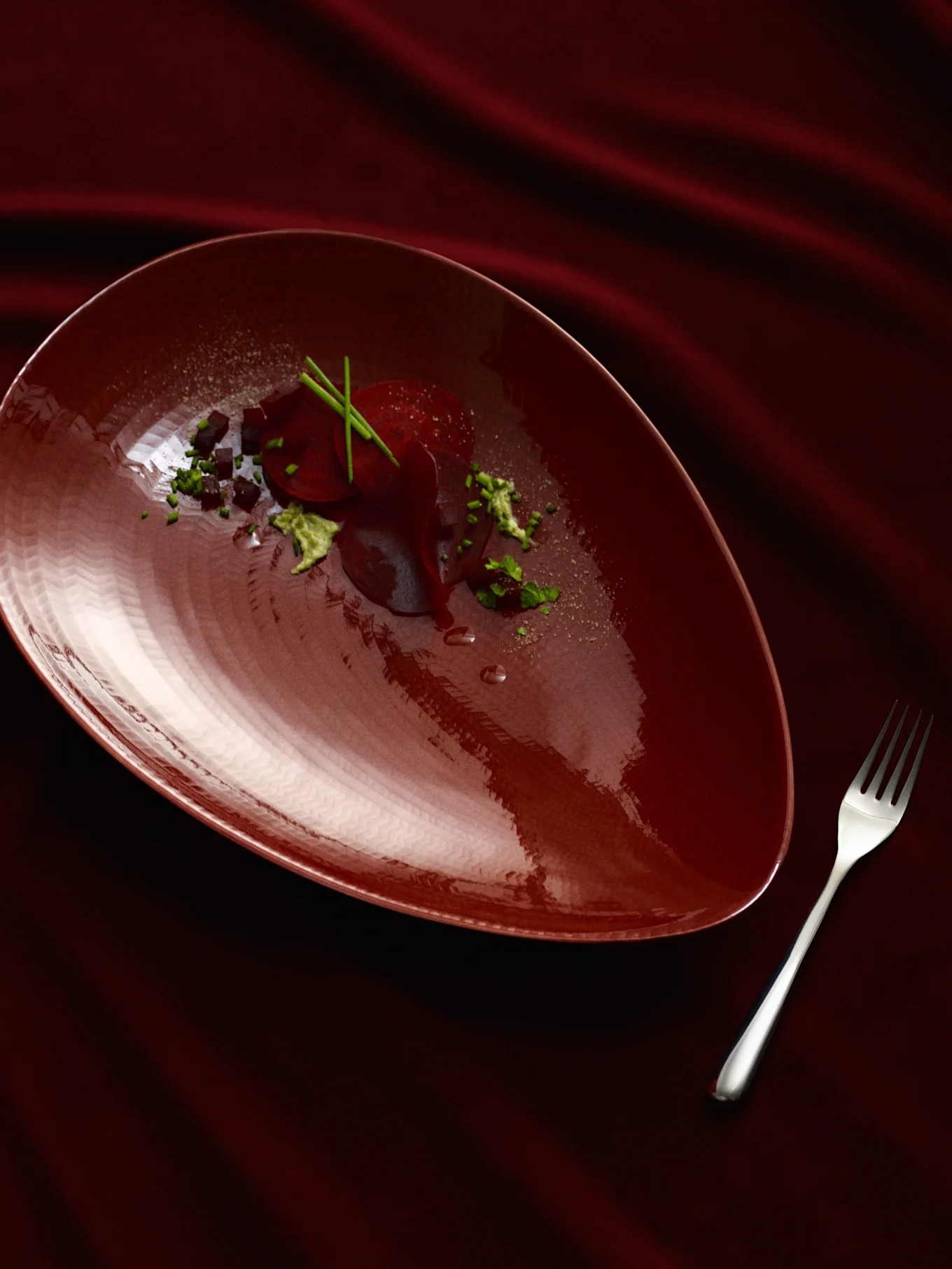 Un piatto a forma di pera rosso scuro con un piatto di barbabietole, guarnito con erbe verdi e cubetti scuri, accanto a una forchetta d'argento su un tessuto rosso scuro drappeggiato.