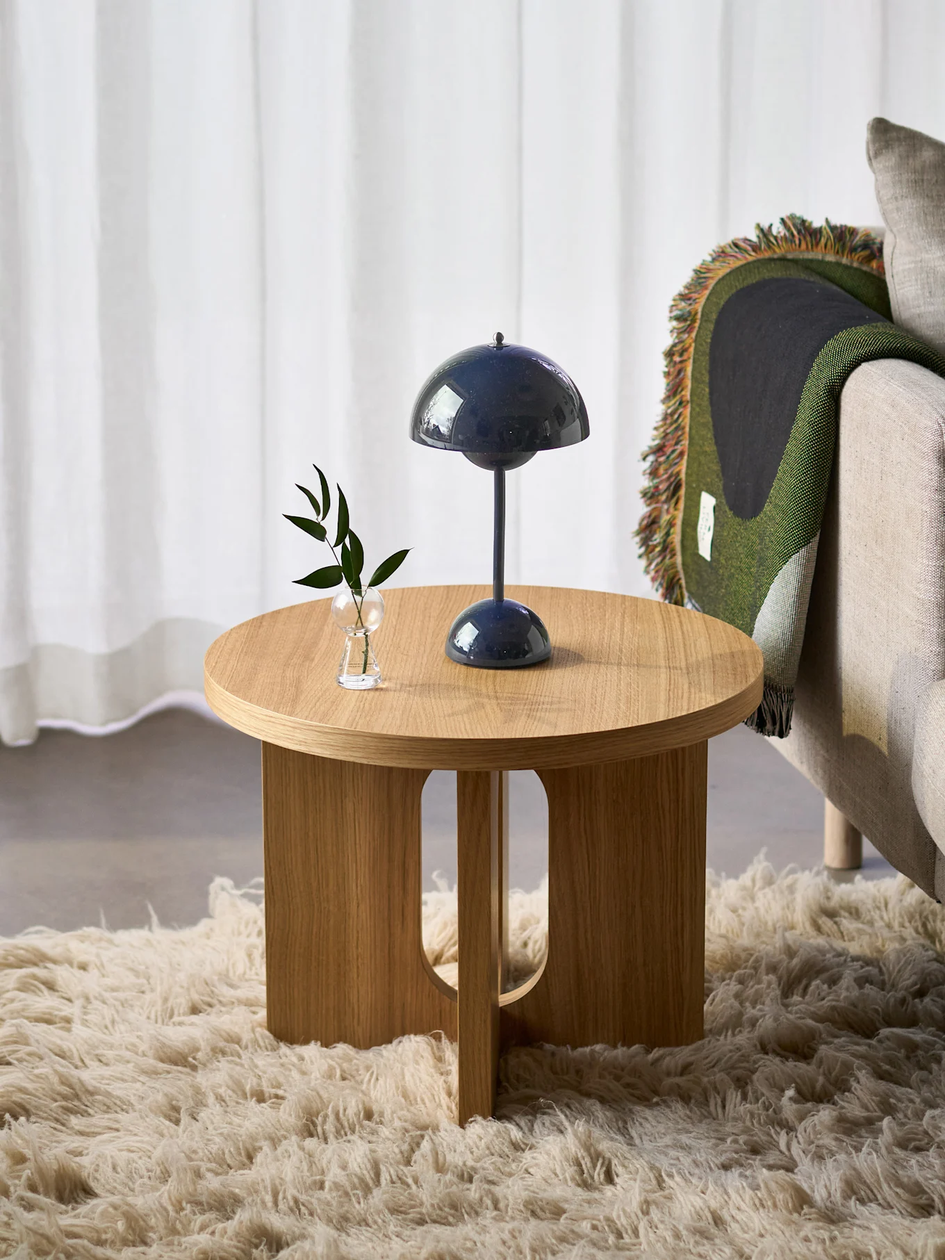 Tavolino rotondo in legno con lampada blu e pianta, accanto a poltrona con coperta verde, su tappeto peloso.