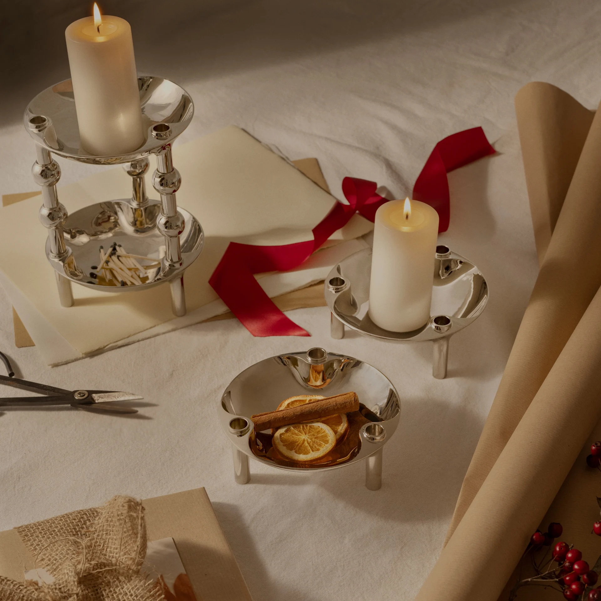 Tre portacandele d'argento, due con candele a pilastro accese, uno con arance essiccate e cannella, disposti con carta da regalo e nastro.