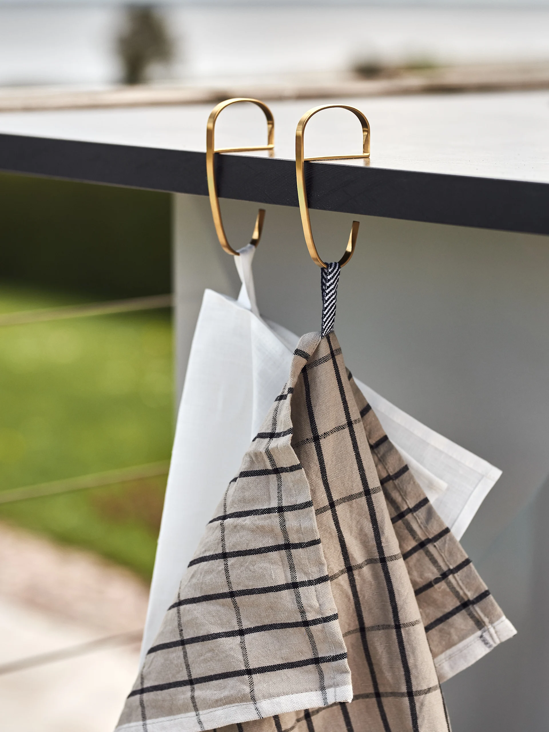 Due strofinacci da cucina, uno bianco e uno a quadri beige, appesi a ganci a S dorati su un bancone scuro.