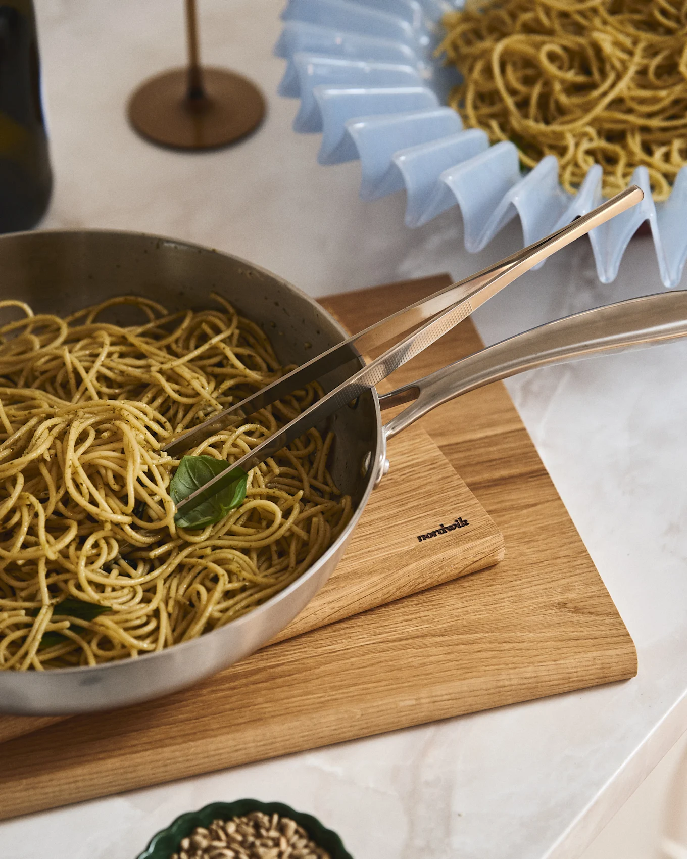 Spaghetti al pesto in padella d'argento con pinze su tagliere in legno Nordwik. Altro piatto di spaghetti in una ciotola azzurra con balze.