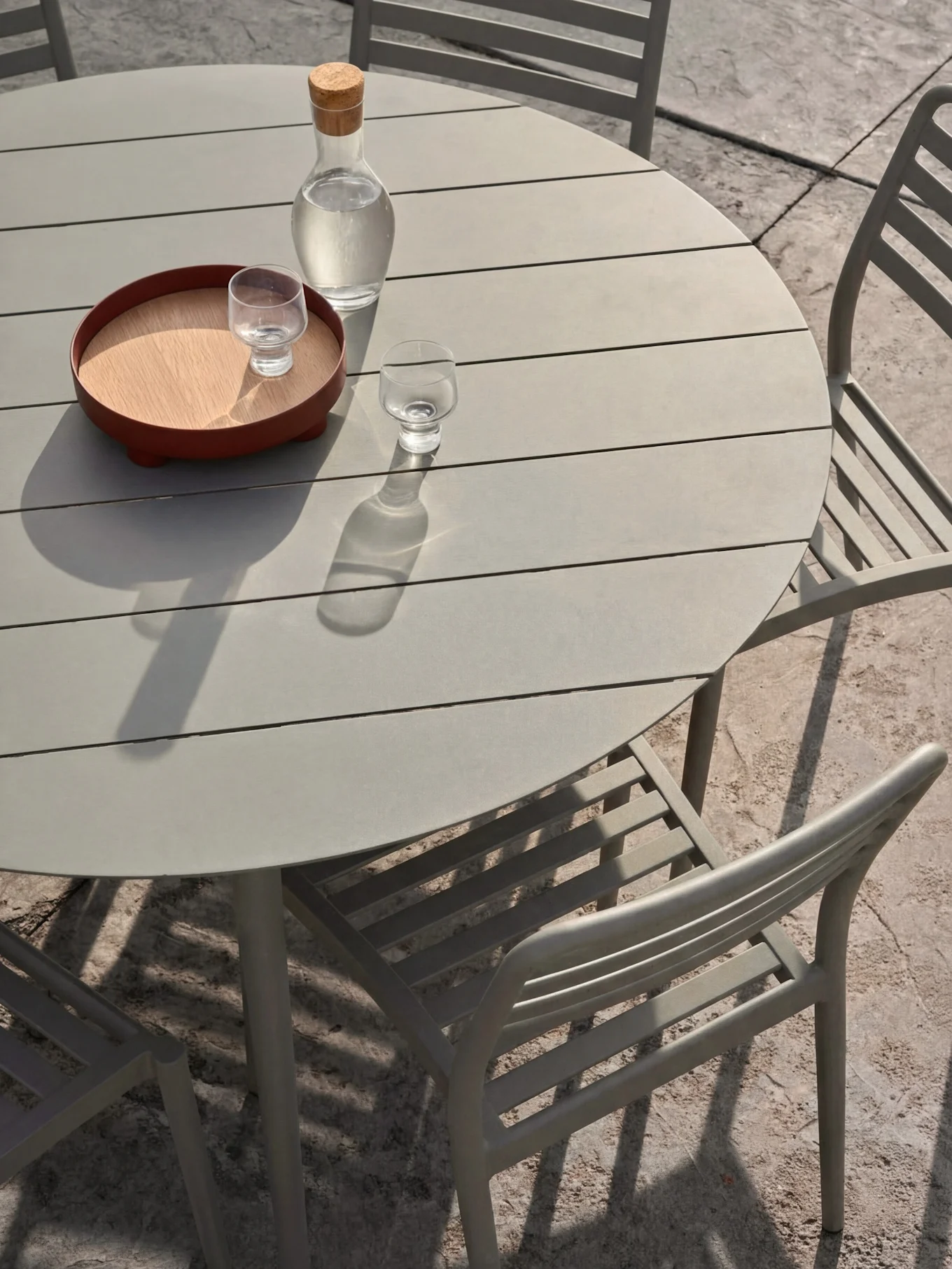 Un set da pranzo all'aperto grigio chiaro con un tavolo rotondo a doghe e sedie. Sul tavolo, una caraffa d'acqua e bicchieri su un vassoio.
