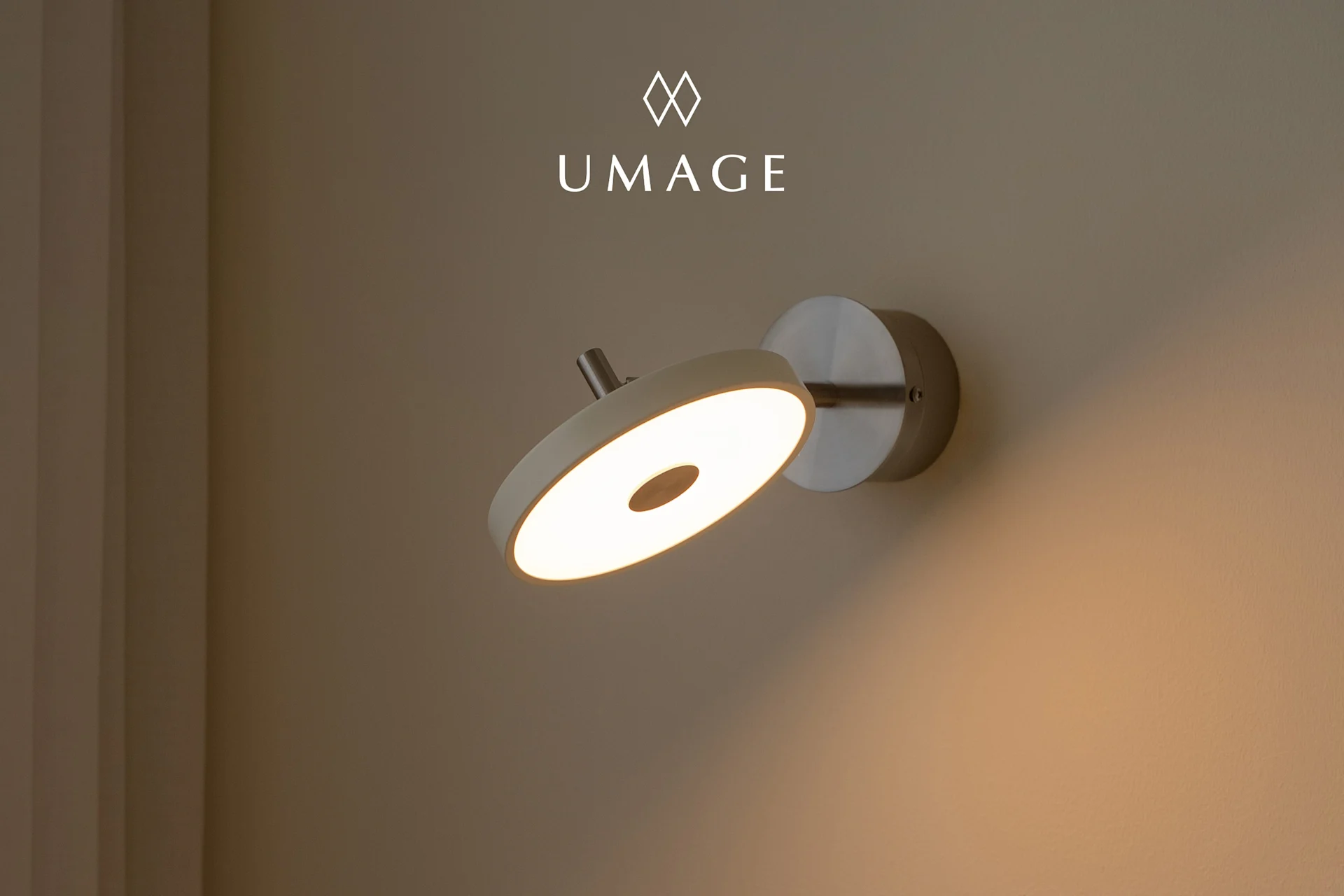 Una lampada da parete circolare moderna con luce calda, montata su una parete marrone chiaro. Logo UMA sopra.