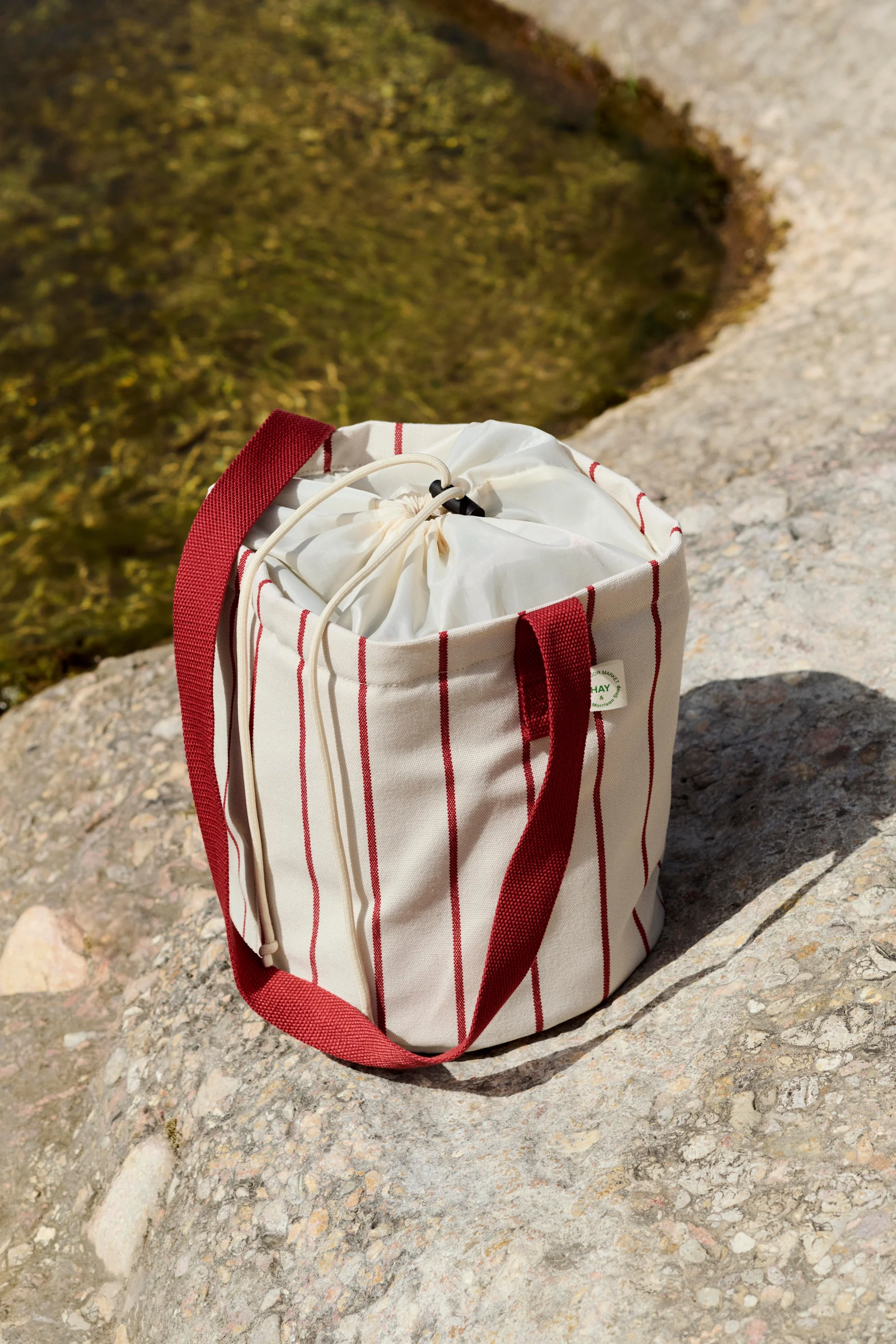 Borsa cilindrica color crema con strisce verticali rosse e chiusura a coulisse, su rocce vicino all'acqua.