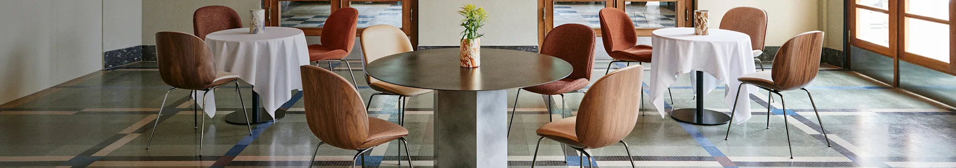 Elegante interno di caffè con un grande tavolo centrale, due tavoli più piccoli con tovaglie bianche e varie sedie su un pavimento a fantasia.