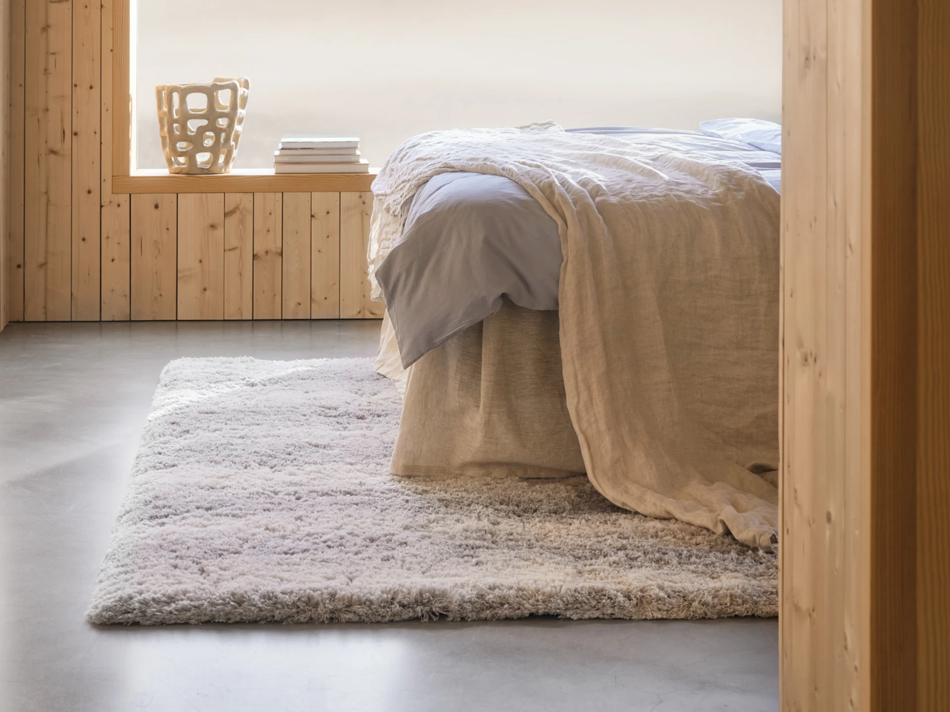 Camera da letto accogliente con letto, lenzuola azzurre, coperta beige, tappeto shaggy, pareti in legno e un vaso decorativo.