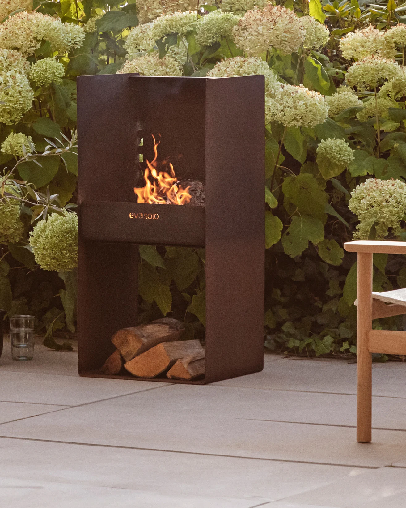 Moderno camino esterno in metallo scuro con fuoco acceso, legna impilata sotto, circondato da piante verdi e un patio.