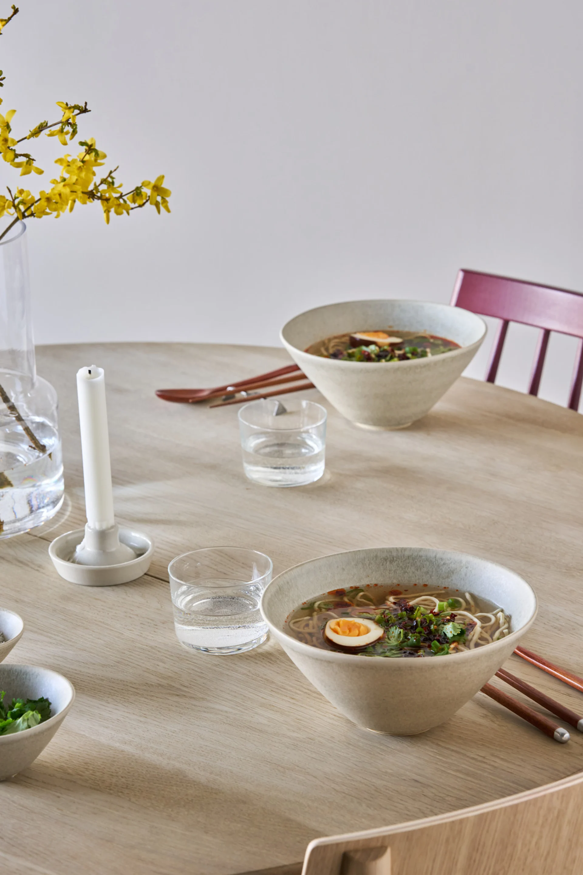 Due ciotole di ramen con uova e guarnizione su un tavolo di legno chiaro, con bicchieri d'acqua e fiori gialli in un vaso.