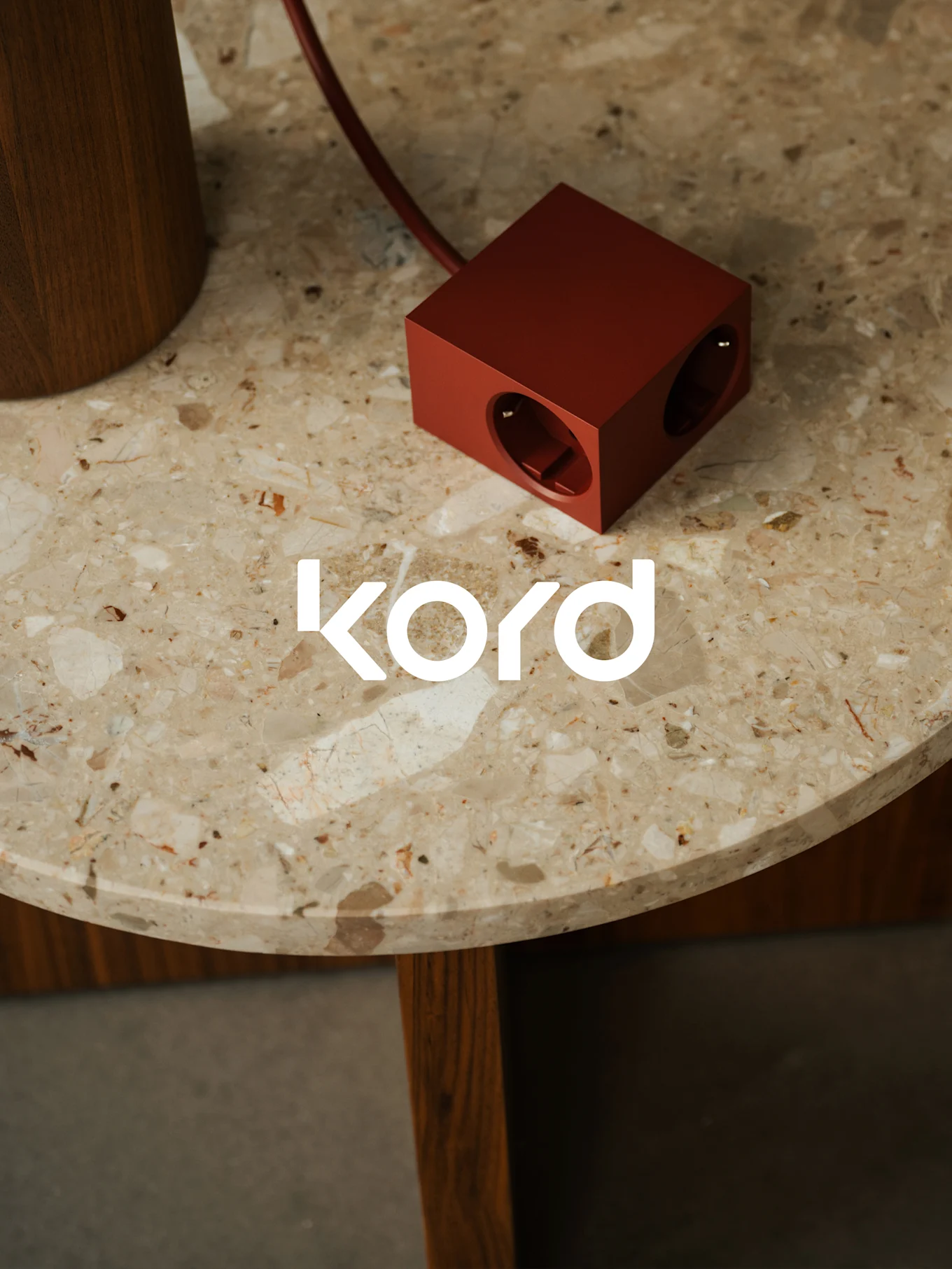 Una ciabatta rosso scuro a forma di cubo con due prese visibili poggia su un tavolo rotondo di marmo, con il logo bianco "kord".