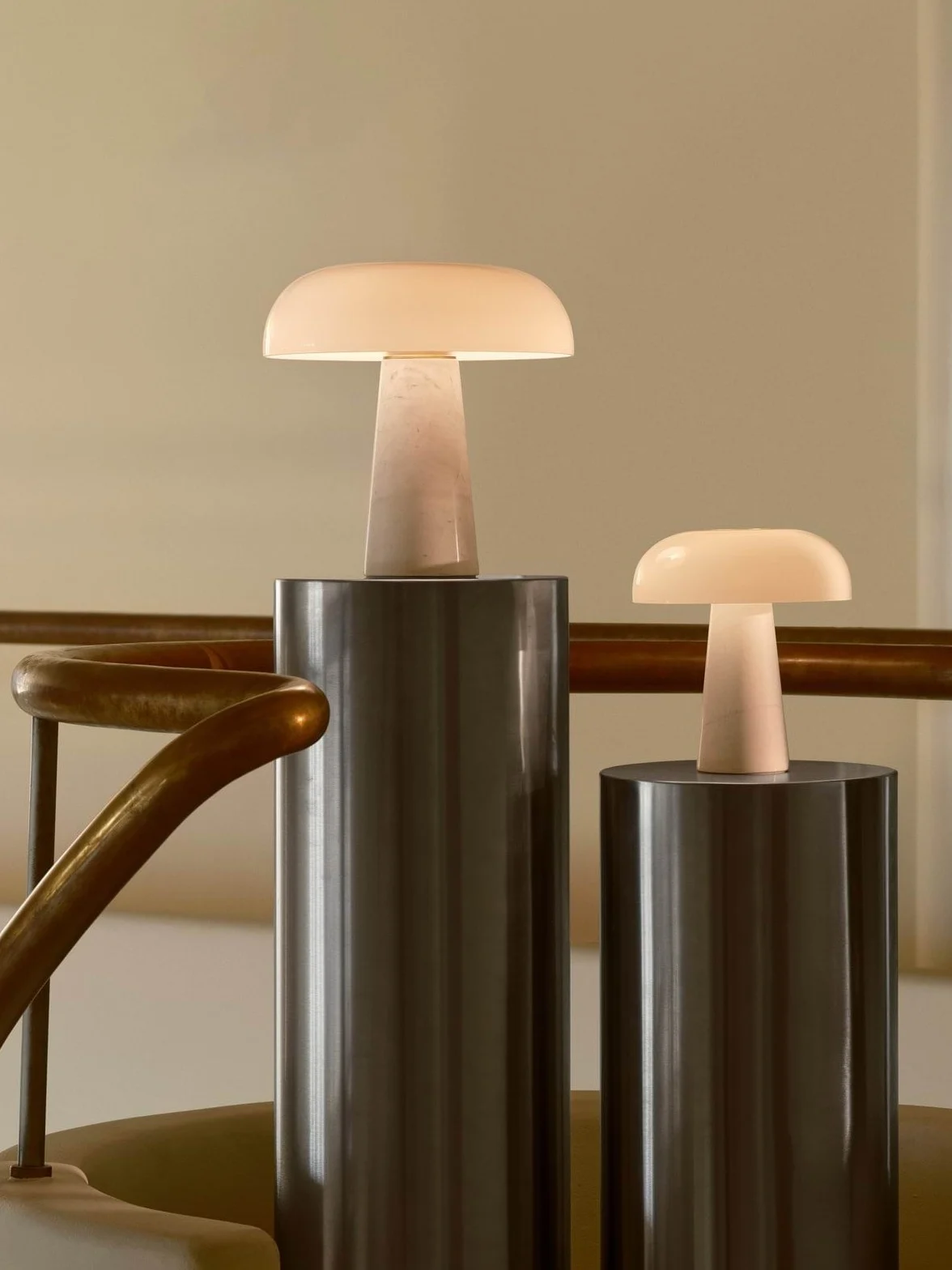 Due lampade da tavolo luminose a forma di fungo, una grande e una piccola, su piedistalli cilindrici accanto a una ringhiera in ottone.