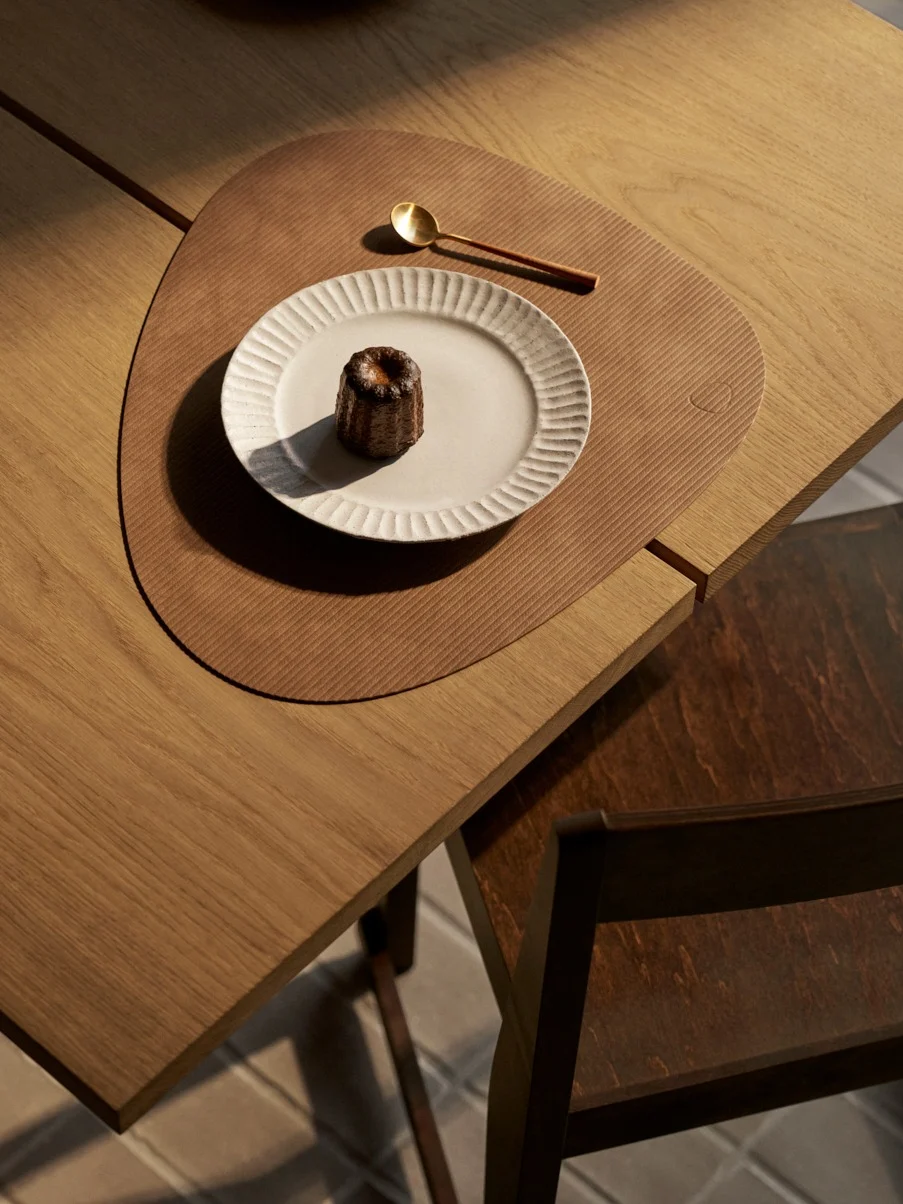 Dall'alto: canelé su piatto bianco, cucchiaio dorato su tovaglietta marrone su tavolo di legno.