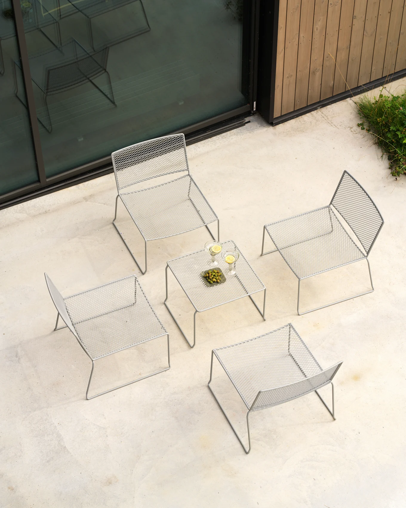 Set lounge esterno grigio chiaro, vista dall'alto: quattro sedie in rete, tavolo con bevande e olive su patio.