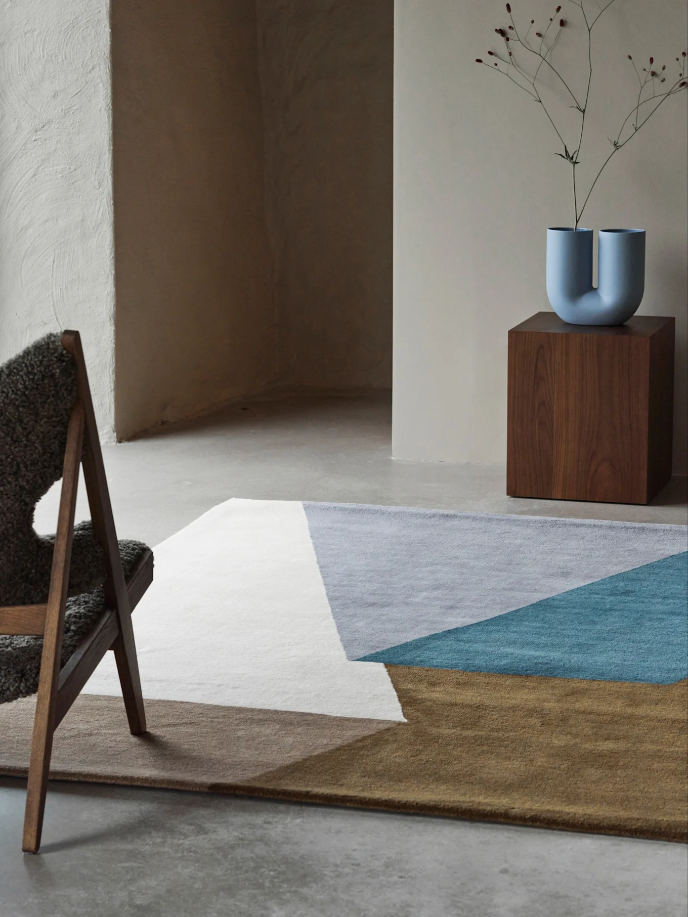 Stanza moderna con un tappeto geometrico bianco, grigio, verde acqua e marrone, un vaso blu a forma di U su un piedistallo di legno e una sedia pelosa.