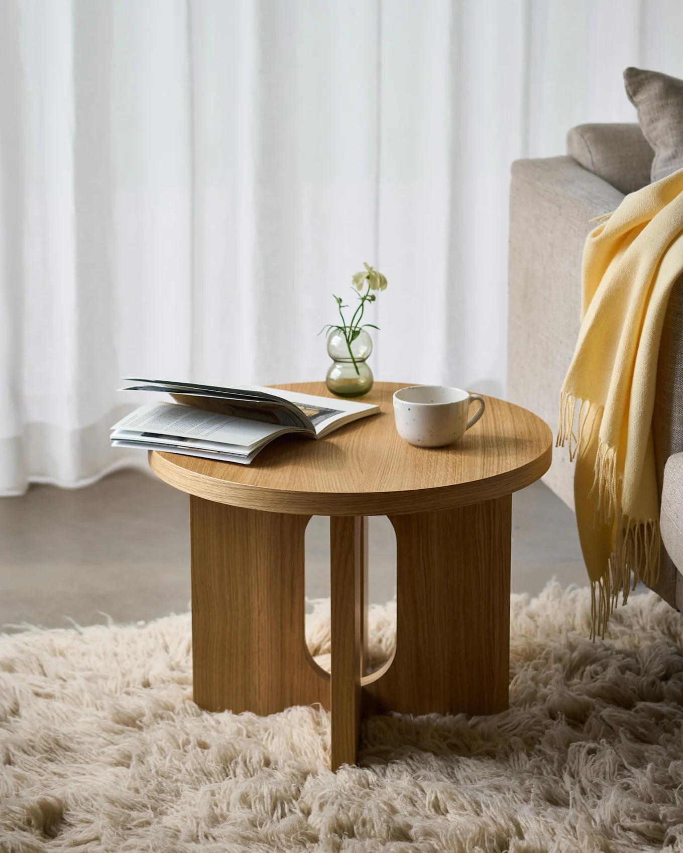 Tavolino rotondo in legno su un tappeto soffice, con una rivista aperta, un fiore bianco in un vaso e una tazza in ceramica, accanto a un divano.