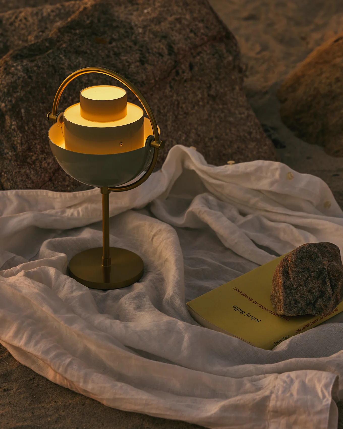 Lampada da tavolo moderna bianca e ottone con luce calda, un libro giallo e una roccia su un panno bianco su superficie rocciosa.