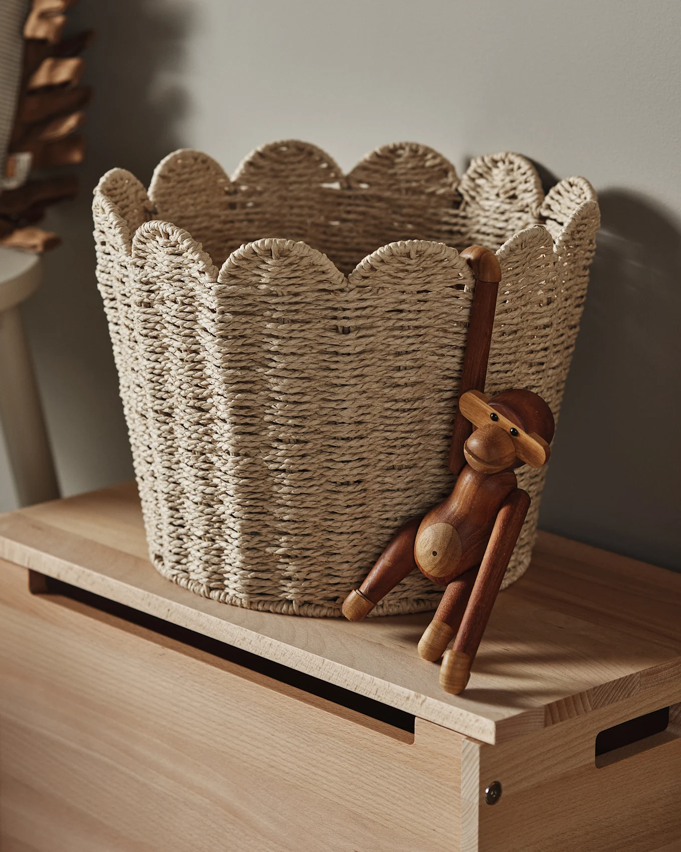 Un cesto intrecciato chiaro con bordo smerlato su una superficie di legno, con una statuetta di scimmia in legno appesa al lato.