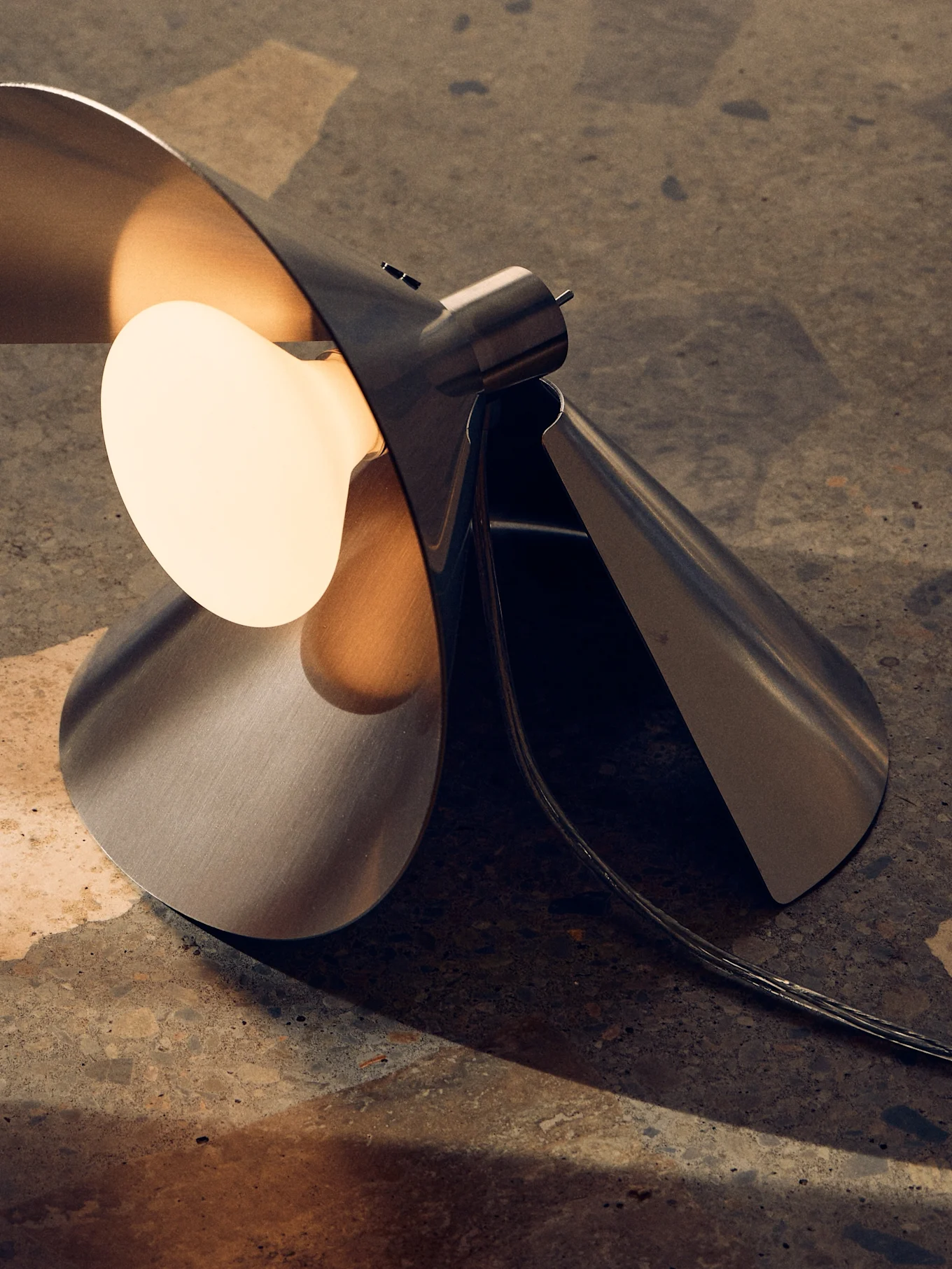 Lampada da tavolo moderna in metallo spazzolato, design scultoreo piegato e lampadina calda esposta su superficie ruvida.