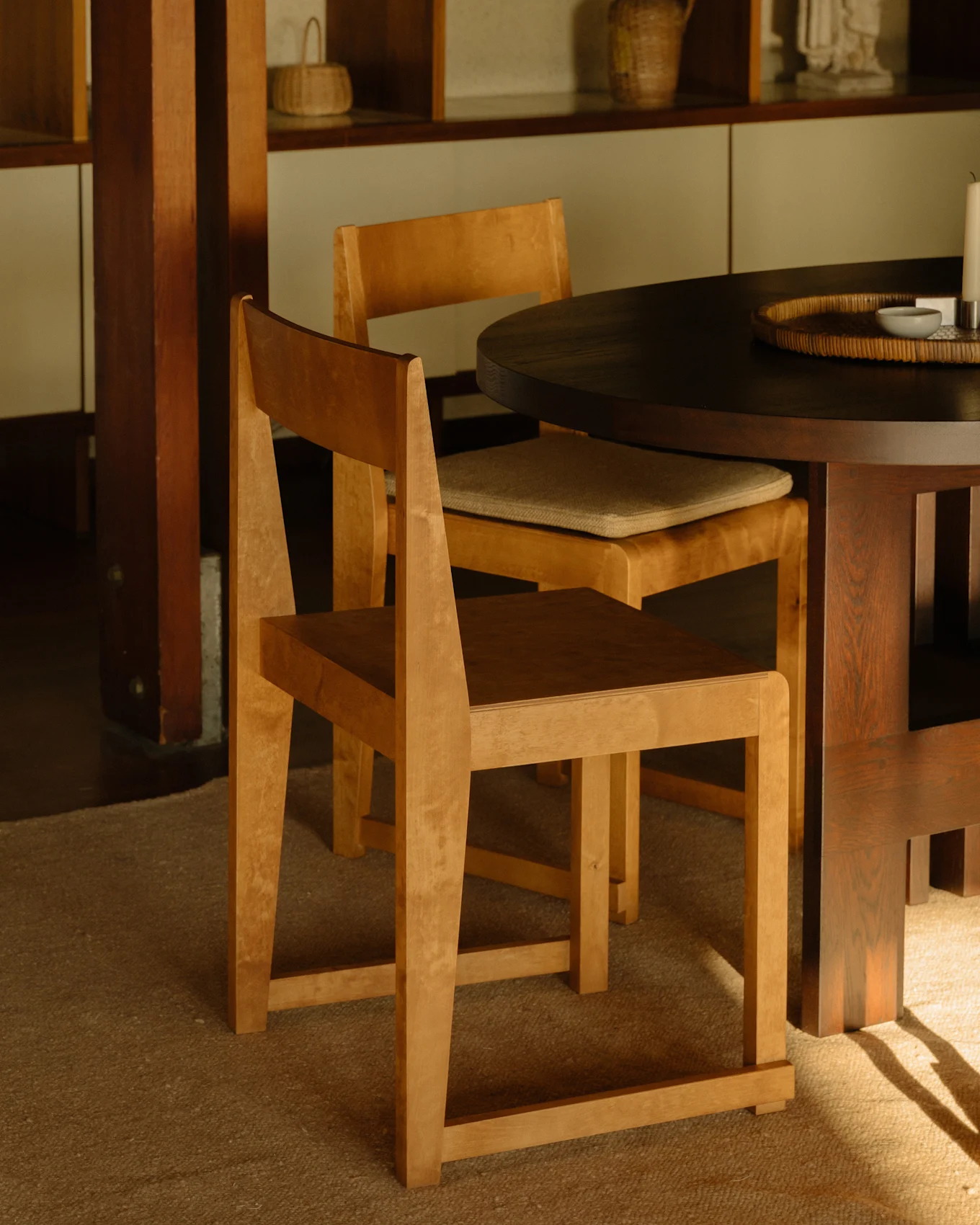 Due sedie in legno chiaro attorno a un tavolo da pranzo rotondo scuro, con scaffali in legno sullo sfondo.