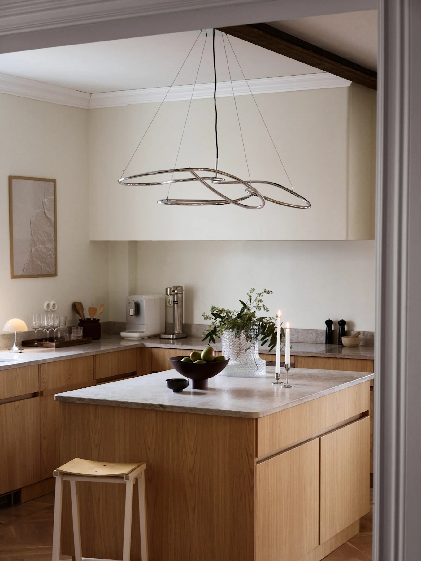 Una cucina moderna con mobili in legno chiaro, ripiani grigi e un lampadario astratto cromato sopra un'isola con frutta e candele.