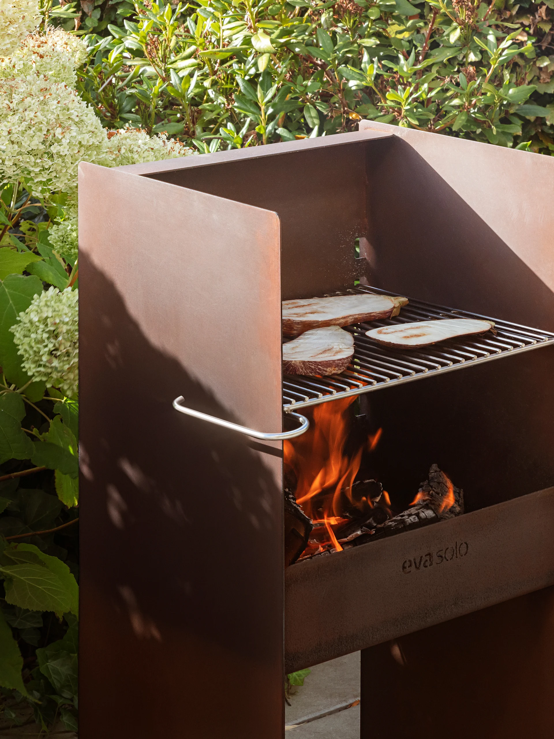 Griglia esterna in metallo arrugginito di Eva Solo con melanzane sulla griglia, fuoco acceso e fogliame lussureggiante.