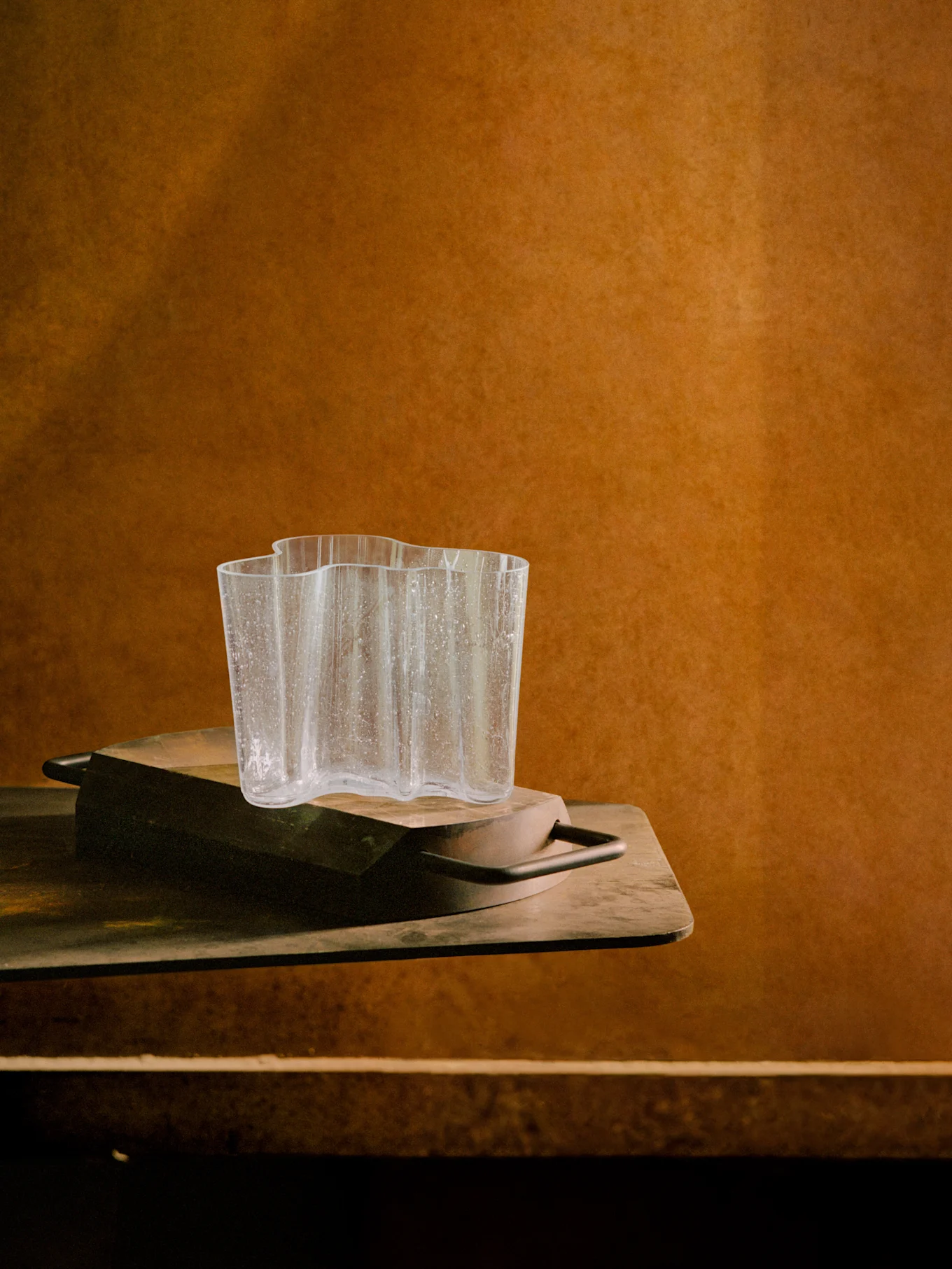 Vaso in vetro trasparente ondulato con goccioline d'acqua su un vassoio di legno scuro, su uno sfondo marrone caldo e testurizzato.