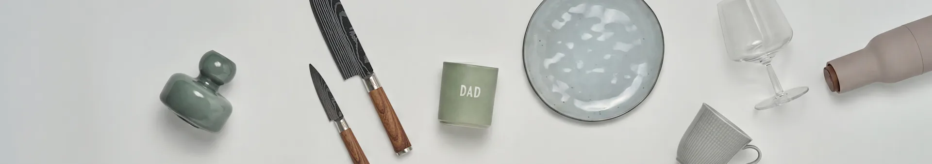 Una disposizione a piatto di articoli da cucina e da regalo: un vaso verde, due coltelli, una candela 'DAD', un piatto in ceramica grigia, un bicchiere da vino, una bottiglia beige e una tazza grigia.