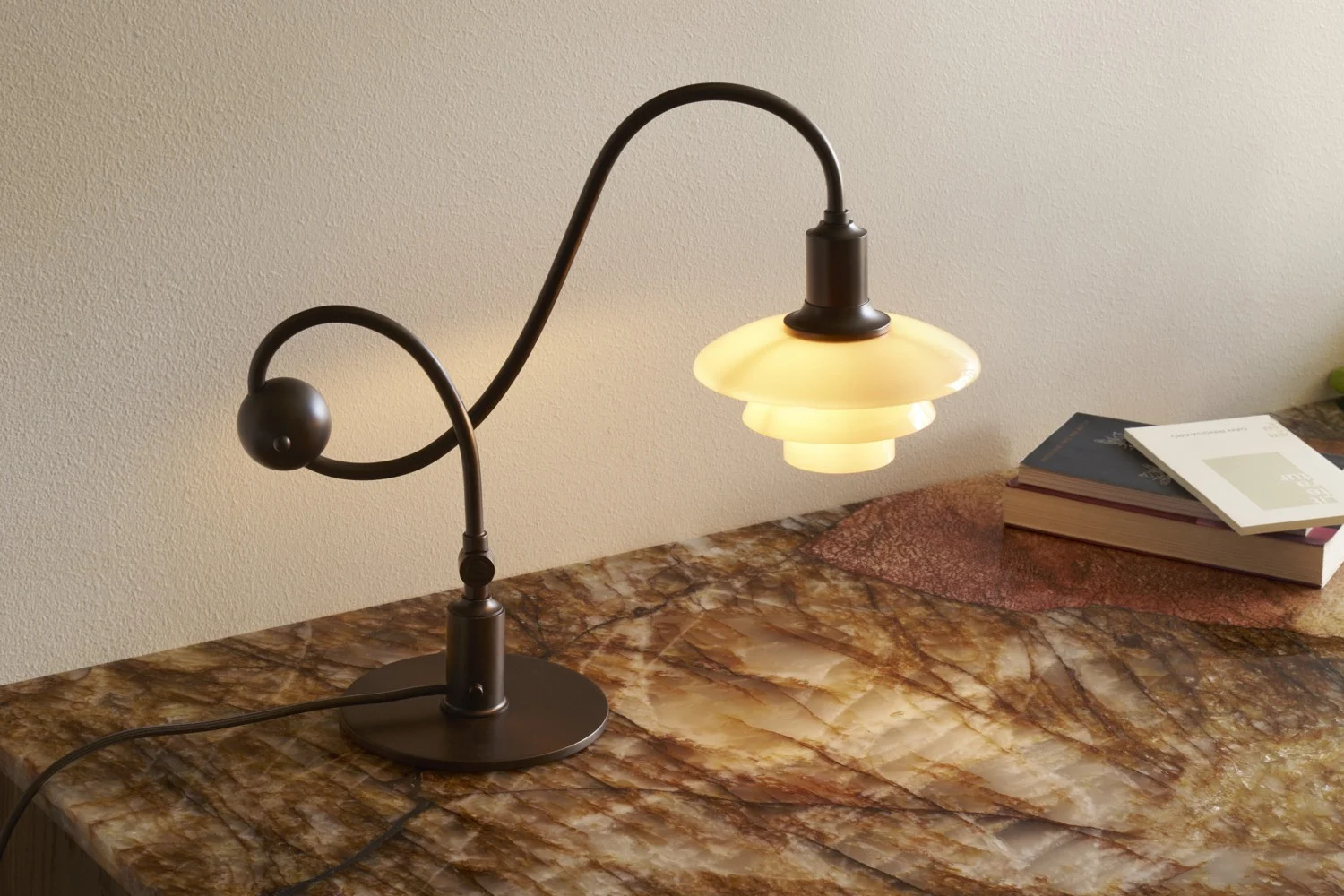 Lampada da tavolo in metallo scuro con stelo curvo e paralume bianco a strati e luminoso su una superficie marmorizzata con libri.