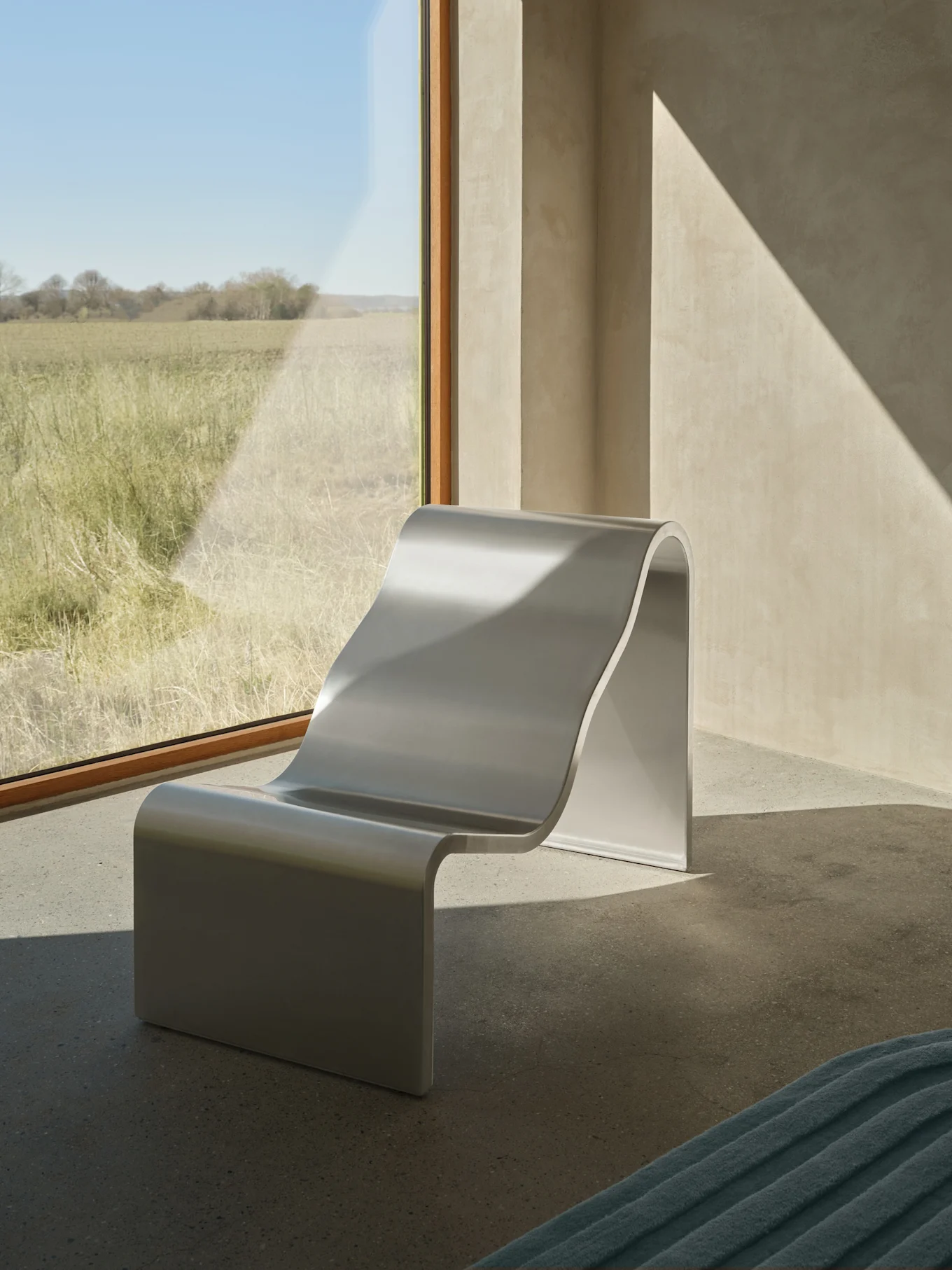 Una moderna poltrona lounge a forma di S color argento si trova accanto a una grande finestra che si affaccia su un campo erboso soleggiato, con motivi di luce solare diagonali sulle pareti.