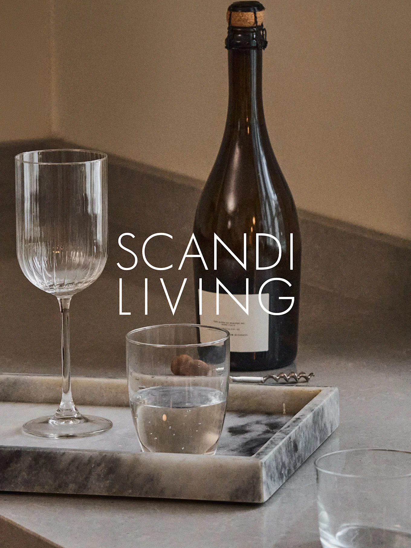 Una bottiglia scura, un calice da vino a coste, un bicchiere d'acqua e un cavatappi su un vassoio di marmo con il testo 'SCANDI LIVING'.