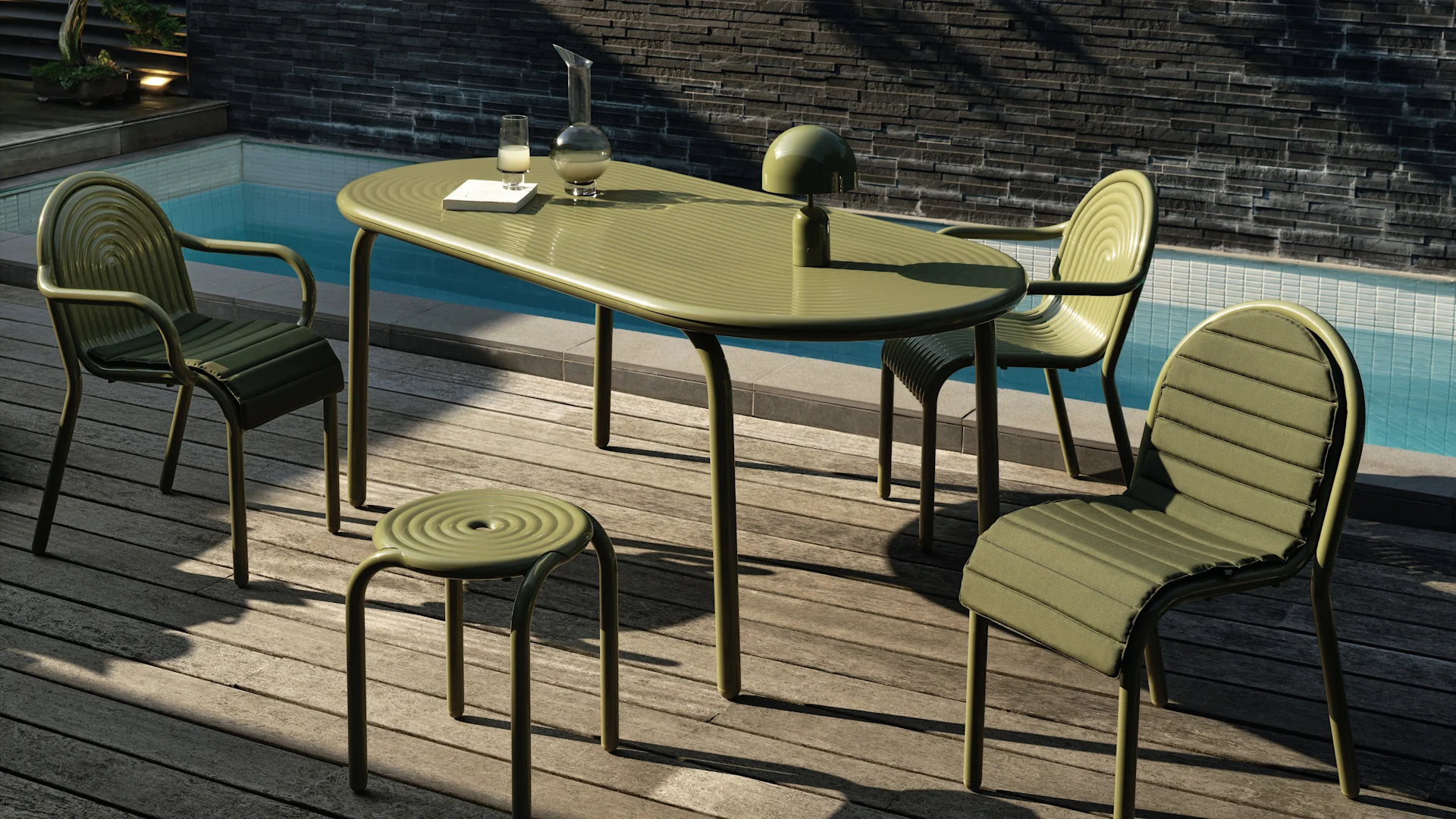 Set da pranzo esterno verde tenue con tavolo ovale, tre sedie imbottite e uno sgabello su un ponte di legno a bordo piscina. Una lampada, caraffa e bicchiere sul tavolo.