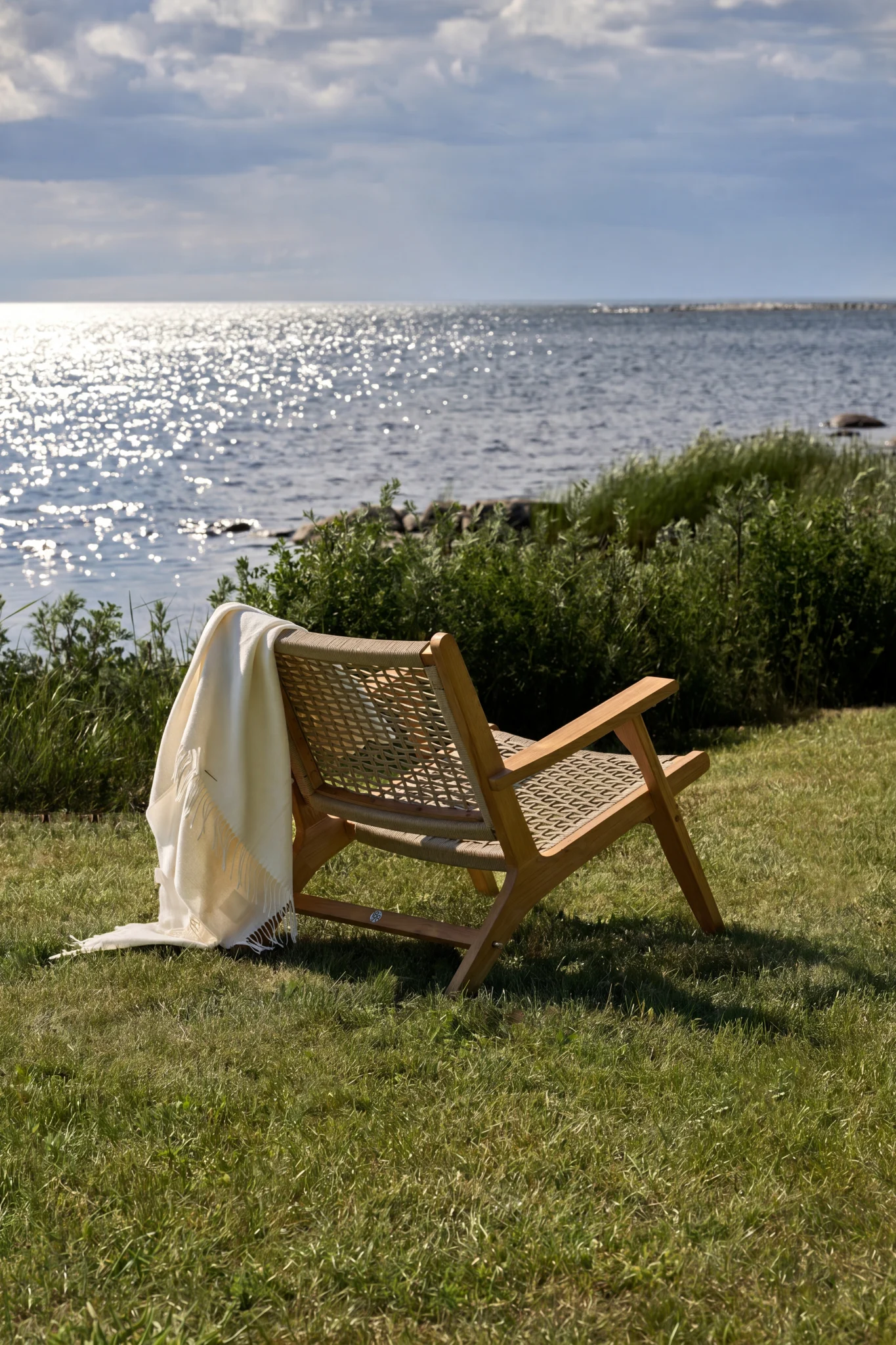 Sedia a sdraio in legno con seduta e schienale intrecciati, drappeggiata con una coperta bianca, di fronte a un oceano scintillante sotto un cielo nuvoloso.