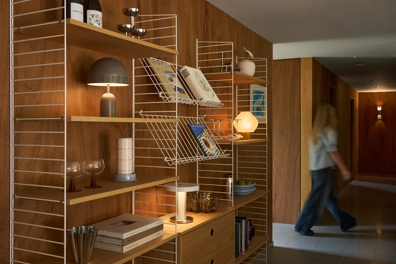 Una persona bionda cammina lungo un corridoio in legno accanto a una moderna libreria piena di libri, lampade e oggetti decorativi.
