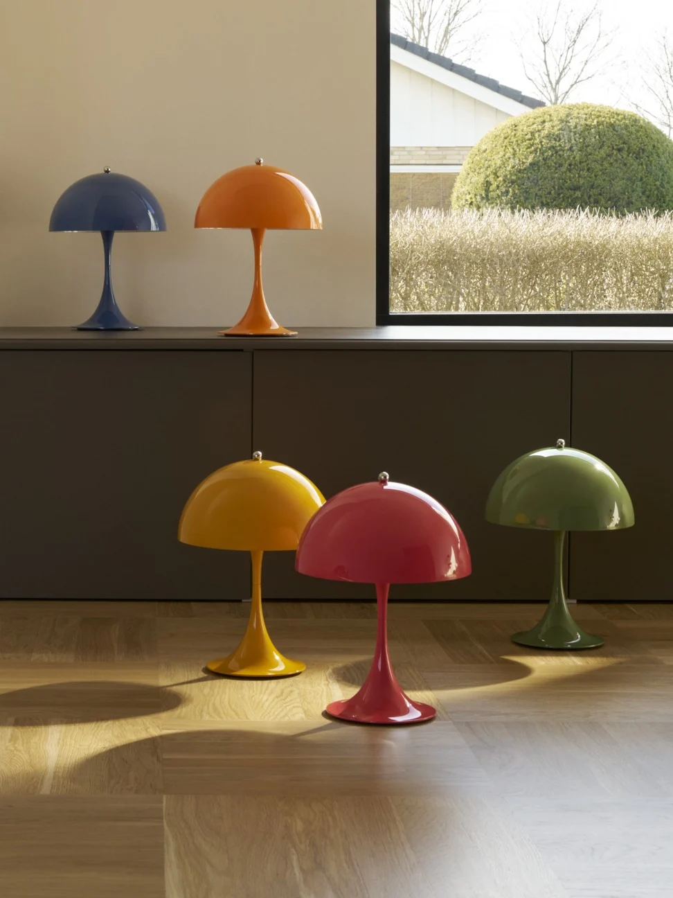 Sei lampade colorate a forma di fungo, due su un mobile e quattro su un pavimento in legno, vicino a una finestra che si affaccia su un giardino.