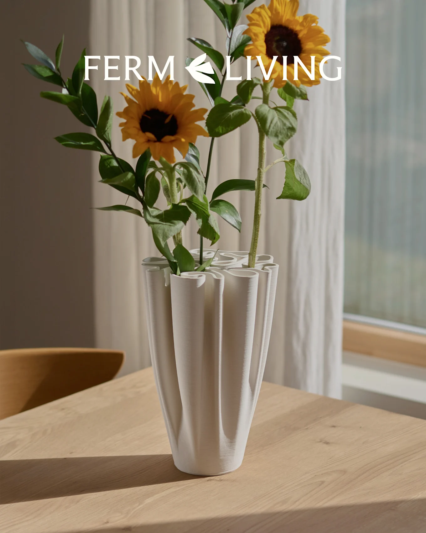 Girasoli in un vaso bianco, a coste e ondulato su un tavolo di legno. Overlay logo Ferm Living.