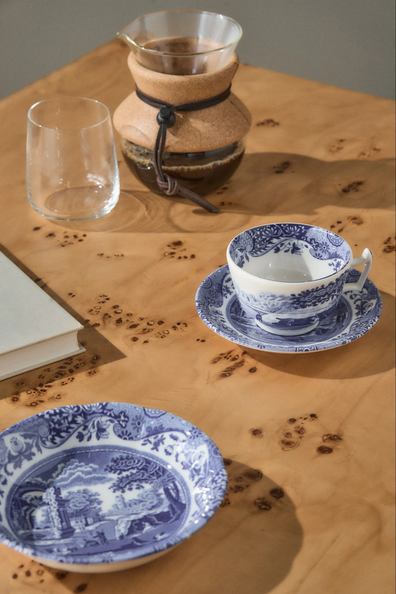 Una caffettiera pour-over, un bicchiere vuoto, una tazza da tè a motivi blu e bianchi su un piattino e un piatto abbinato su un tavolo di legno chiaro.