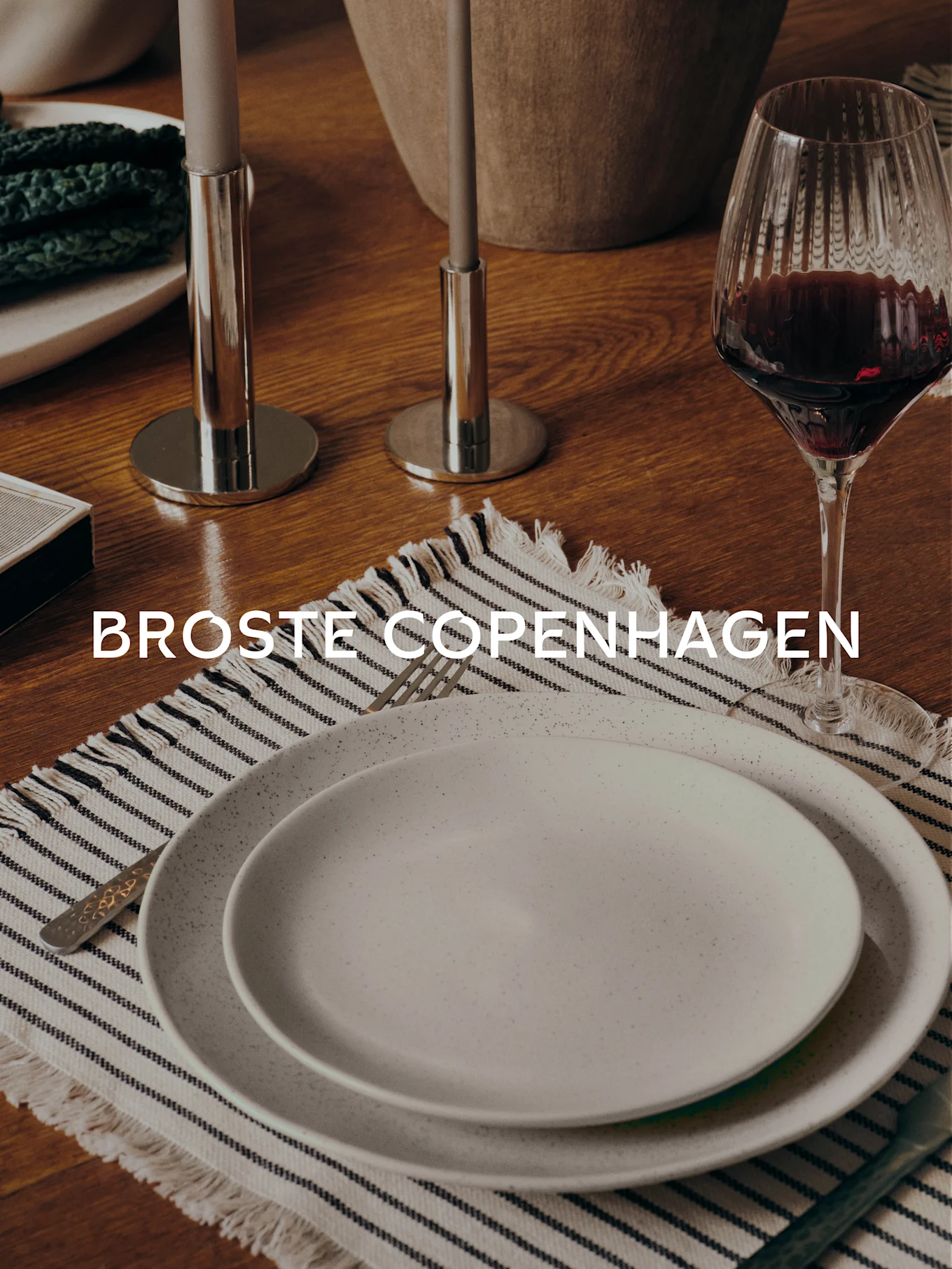 Tavolo da pranzo con due piatti maculati su una tovaglietta a righe, forchetta, bicchiere di vino, due candelabri e la scritta 'BROSTE COPENHAGEN'.