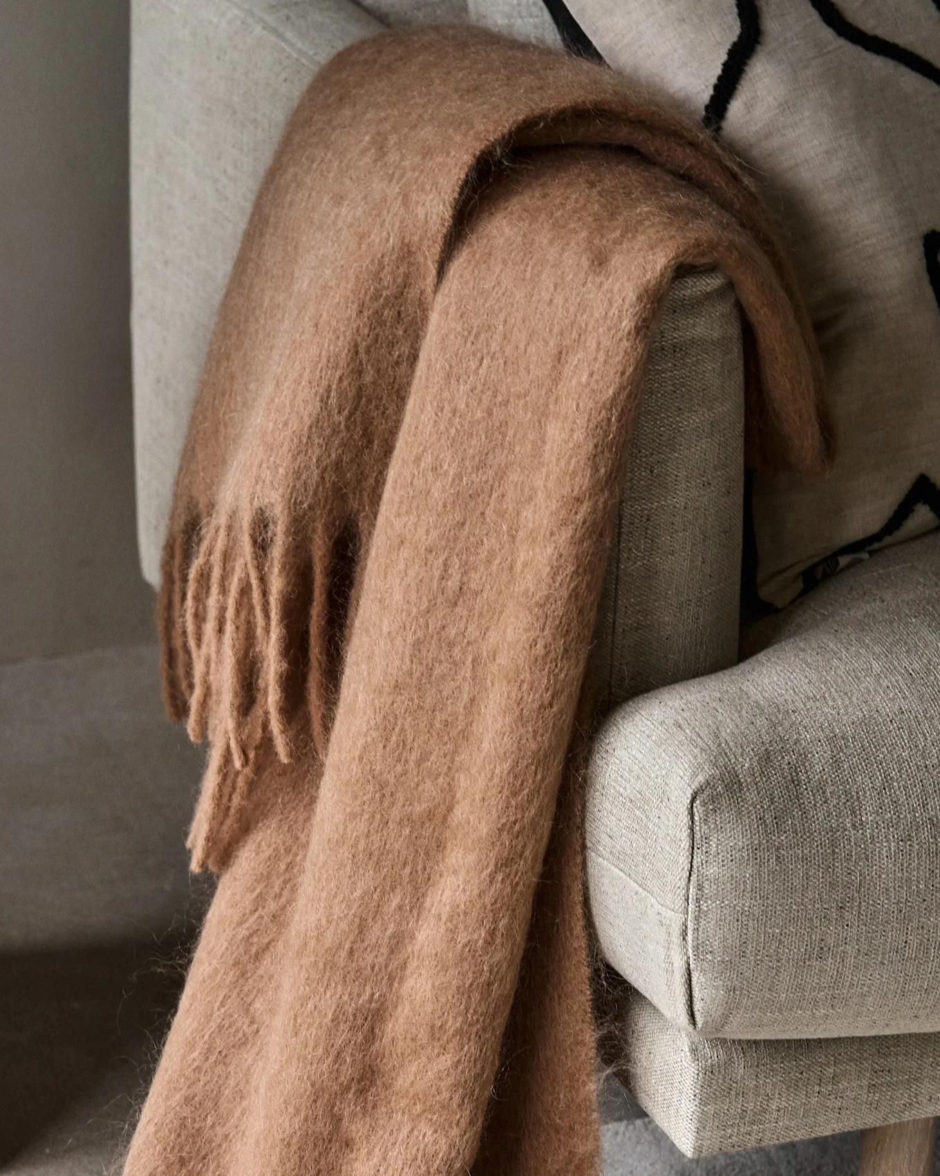 Plaid soffice marrone chiaro con frange, drappeggiato su una poltrona grigio chiaro.