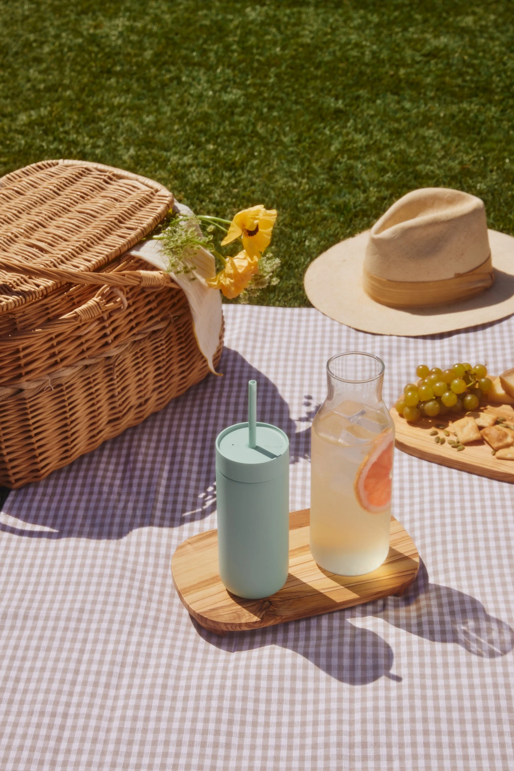 Picnic su una coperta a quadri: cesto di vimini, bicchiere verde chiaro, bevanda al pompelmo, uva e cracker sull'erba.