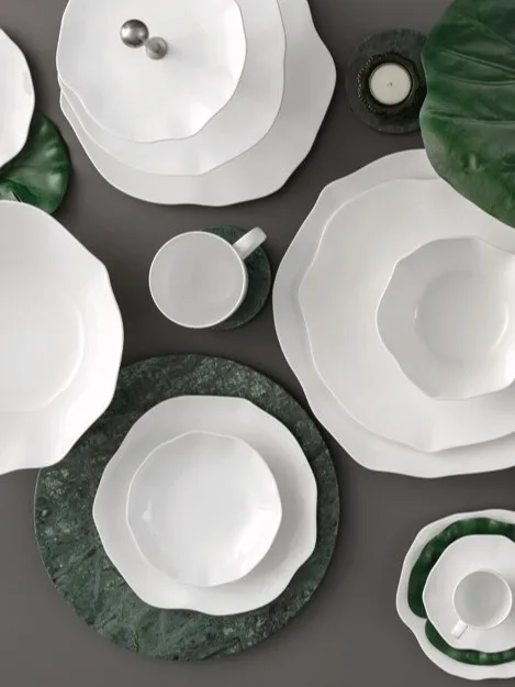Una collezione di stoviglie in ceramica bianca con bordi ondulati, disposta con foglie verdi e sottobicchieri in marmo su una superficie scura.