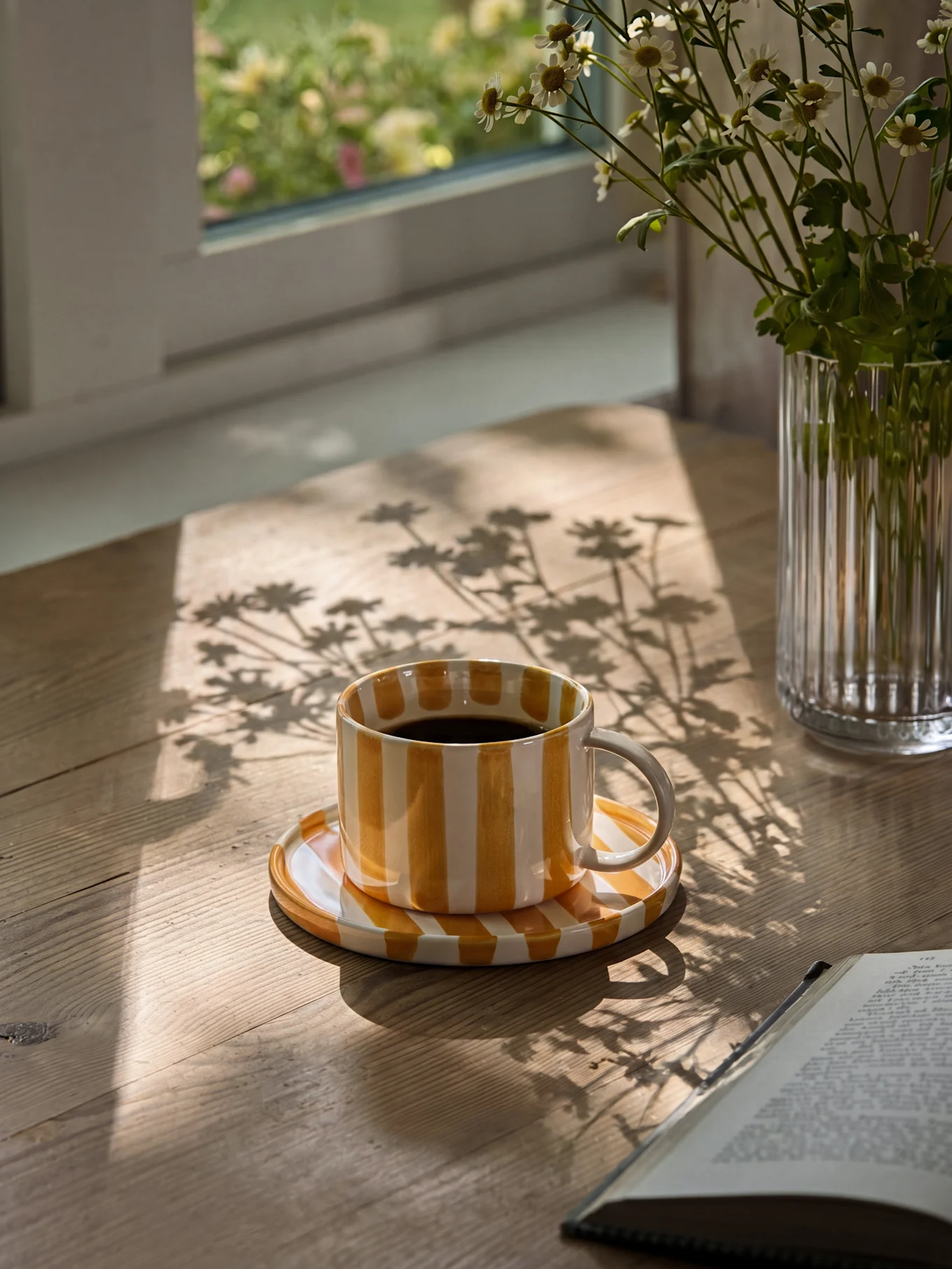 Tazza da caffè a righe gialle e bianche con caffè nero su un tavolo di legno con ombre del sole e un vaso di margherite.