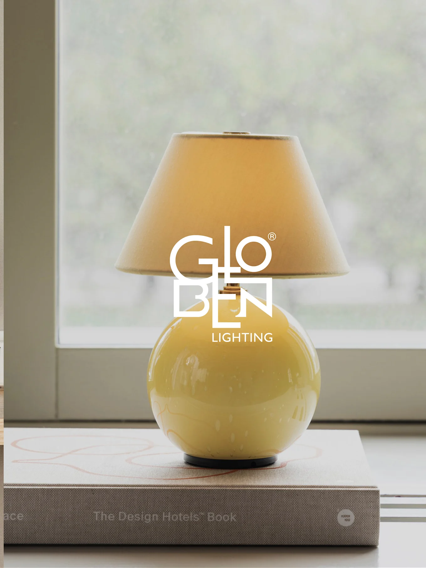 Lampada da tavolo gialla con paralume beige, appoggiata su libri vicino a una finestra.
