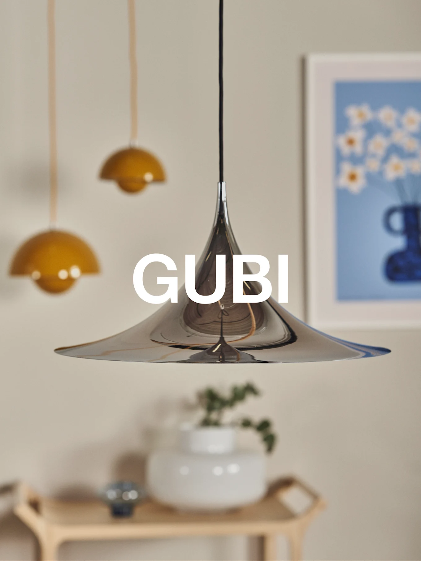 Una lampada a sospensione conica in cromo lucido con il logo GUBI, su sfondo sfocato con altre lampade e opere d'arte.